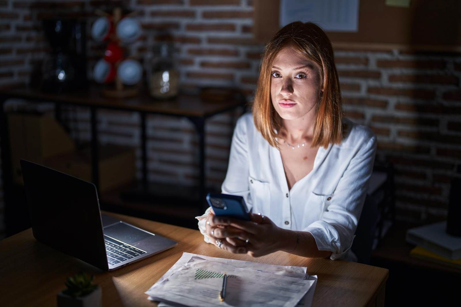 Mulher sentada em uma mesa de escritório à noite, segurando um smartphone e com um notebook e documentos financeiros à sua frente, ilustrando a gestão digital da portabilidade de crédito.