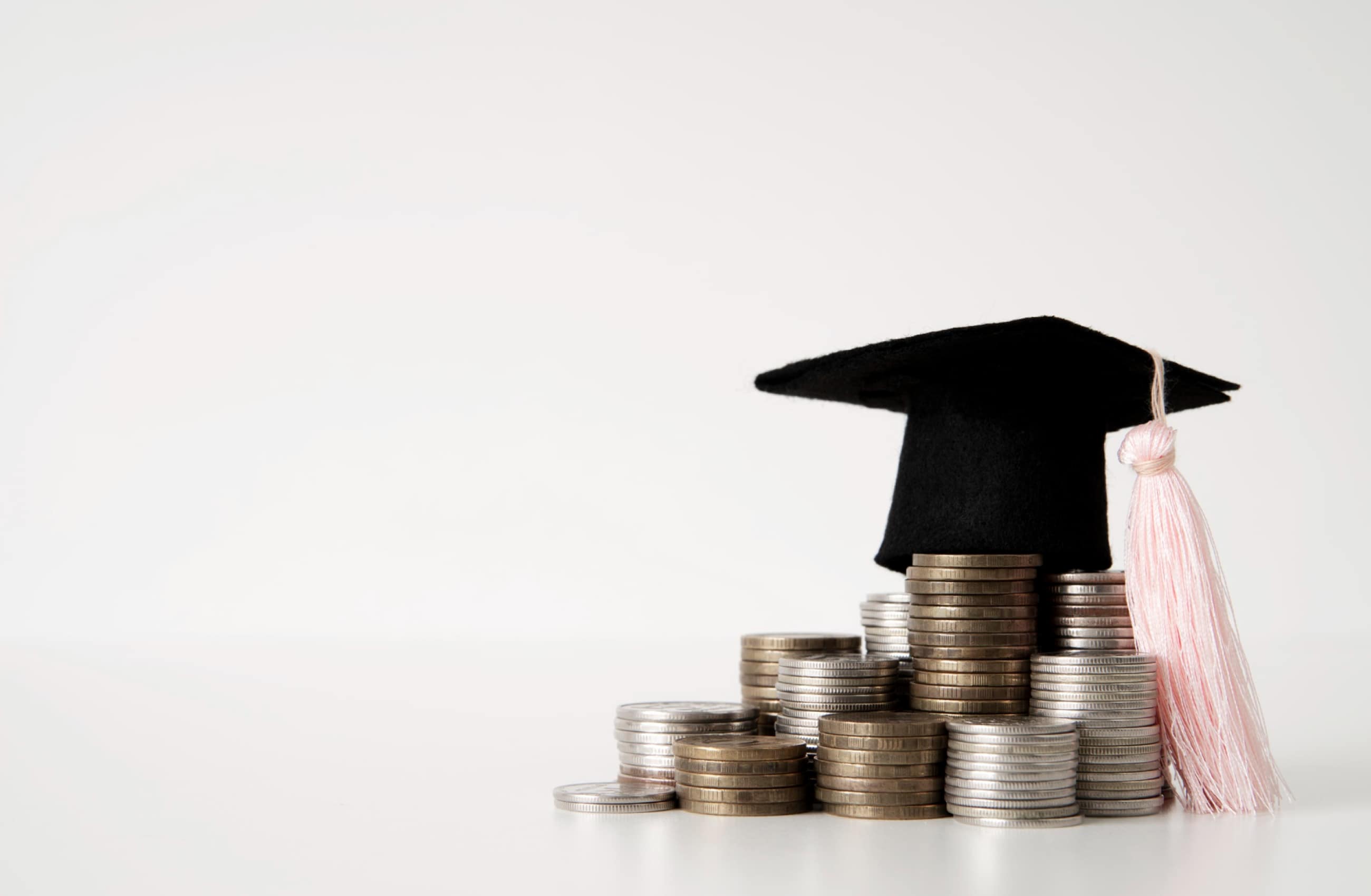 Mini capelo de formatura preto sobre pilhas de moedas em fundo branco, simbolizando o investimento para fazer pós-graduação.
