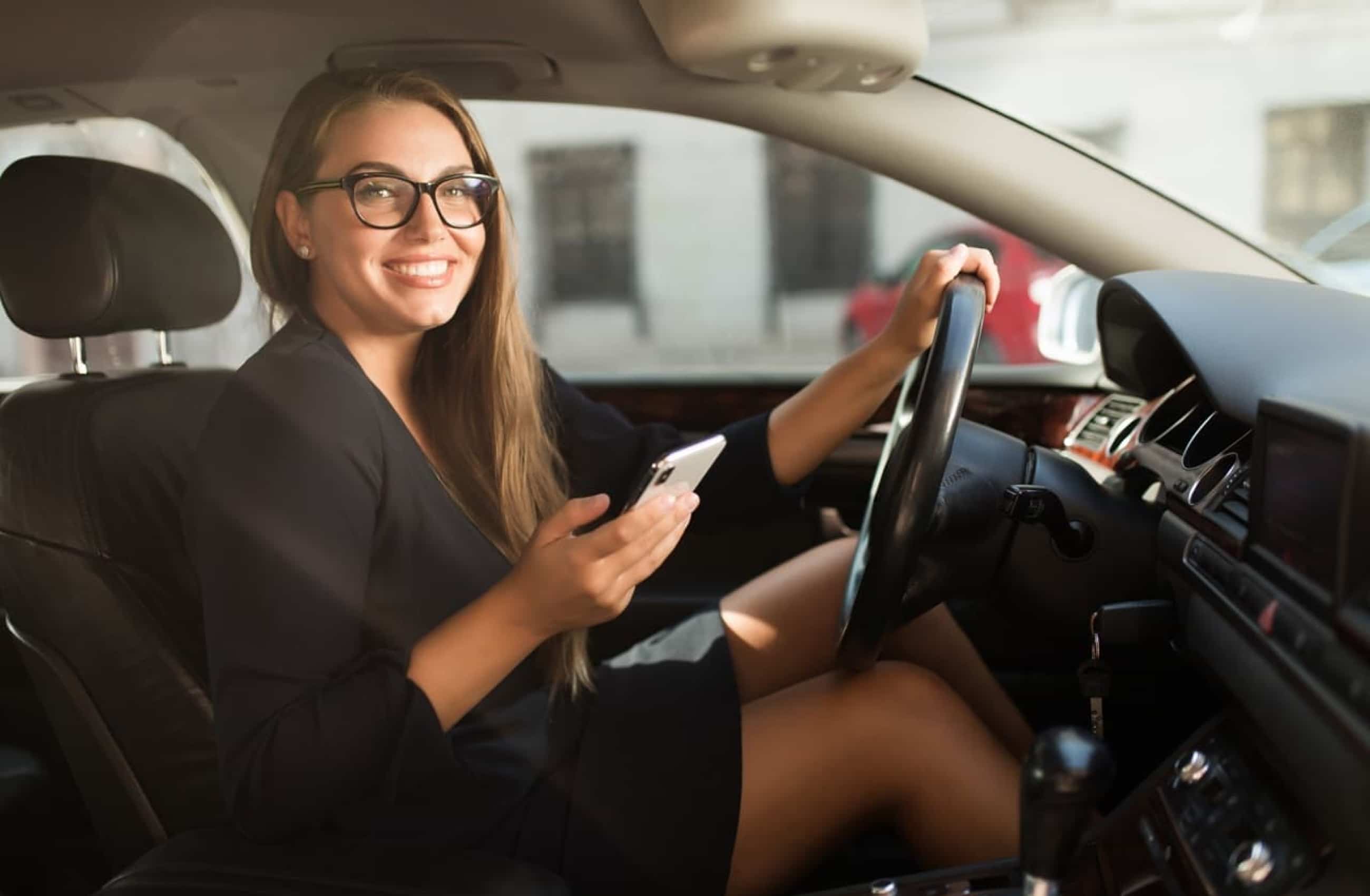 Mulher branca está usando um vestido preto e óculos de grau, sentada no banco do motorista de um carro. Enquanto uma das mãos está no volante, a outra segura um celular. Ela está olhando para a câmera e sorrindo.