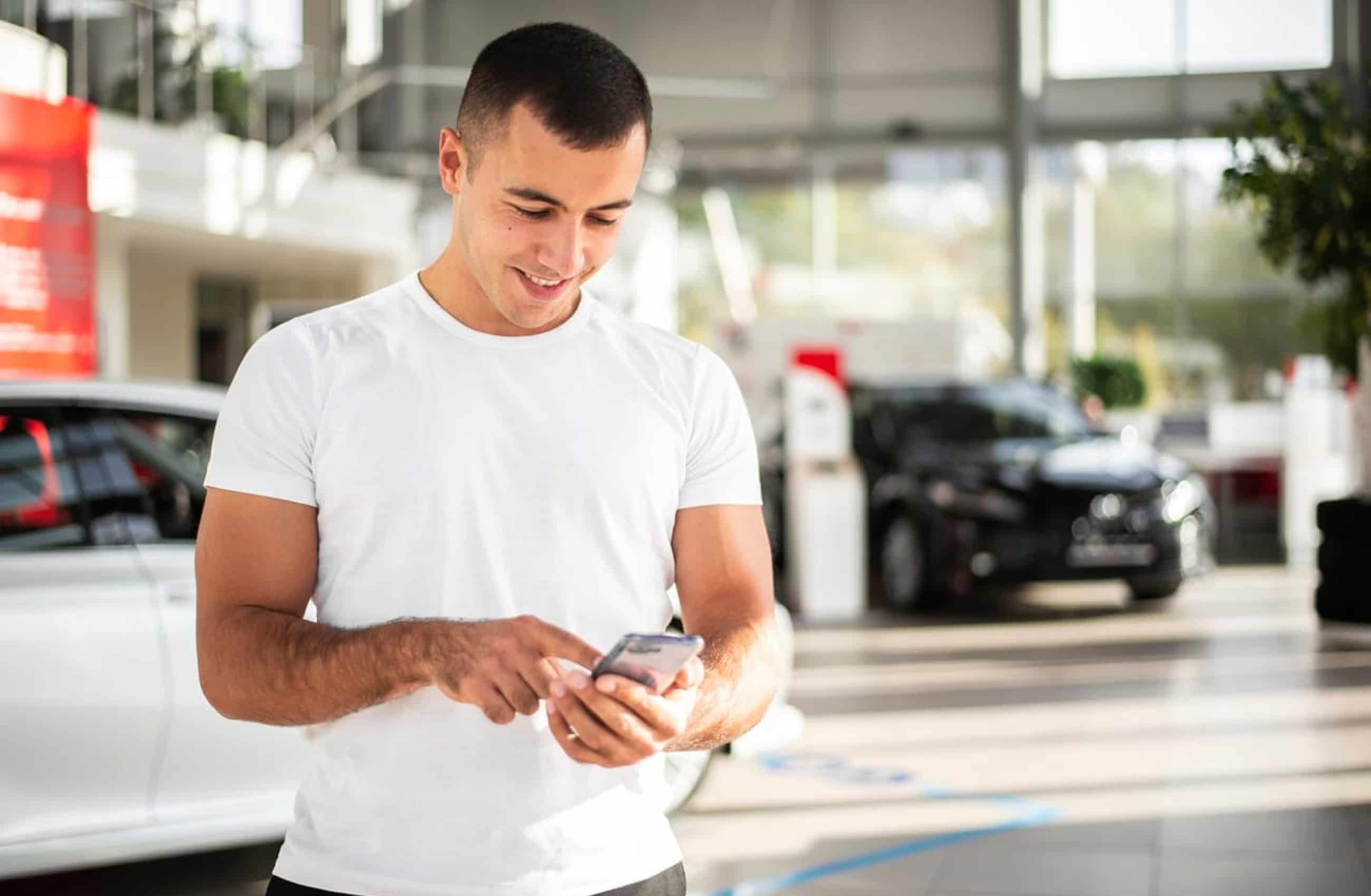 Homem branco vestindo uma camiseta branca, está sorrindo enquanto mexe no celular. Ao fundo desfocado, tem dois carros dentro de uma concessionária.