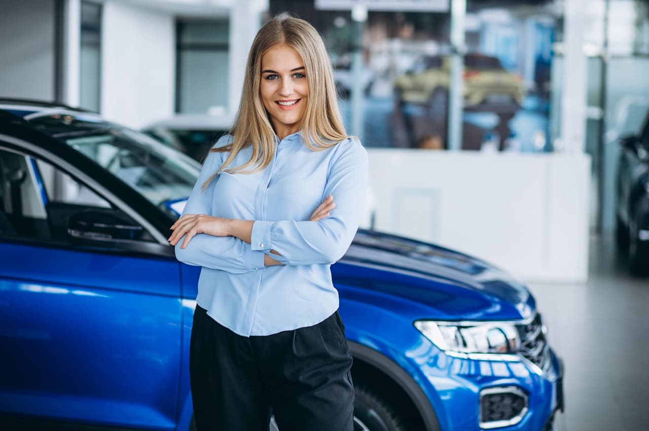 Mulher loira sorridente, vestindo camisa azul, com os braços cruzados na frente de um veículo azul em uma concessionária.