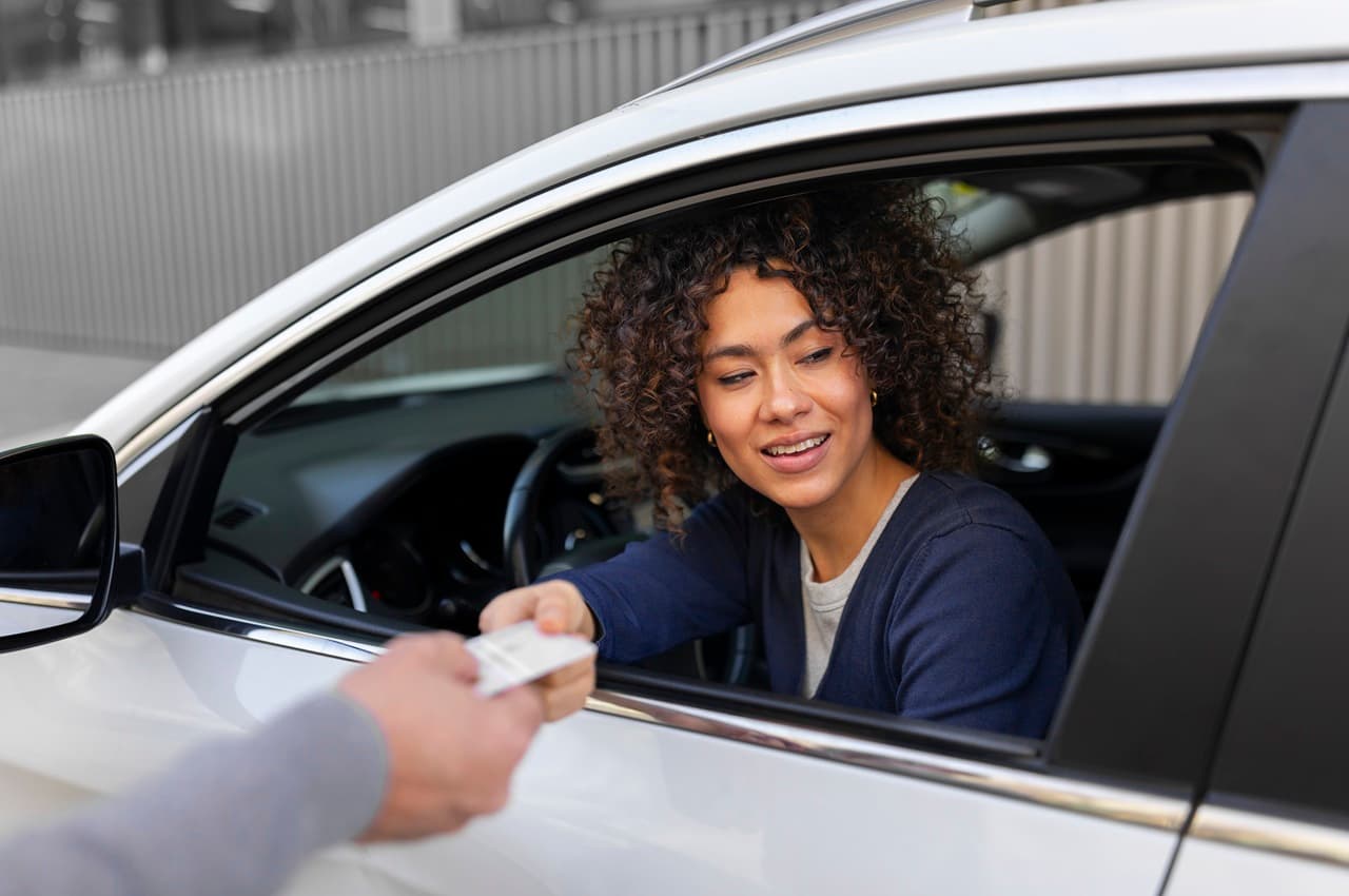 Mulher de cabelo cacheado sorrindo enquanto recebe a sua CNH de dentro de um carro.