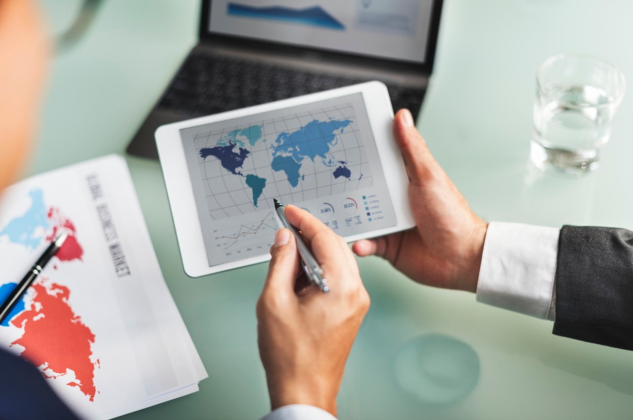 Pessoa segurando um tablet exibindo um mapa-múndi com gráficos de dados, sugerindo planejamento de operações onshore e offshore.