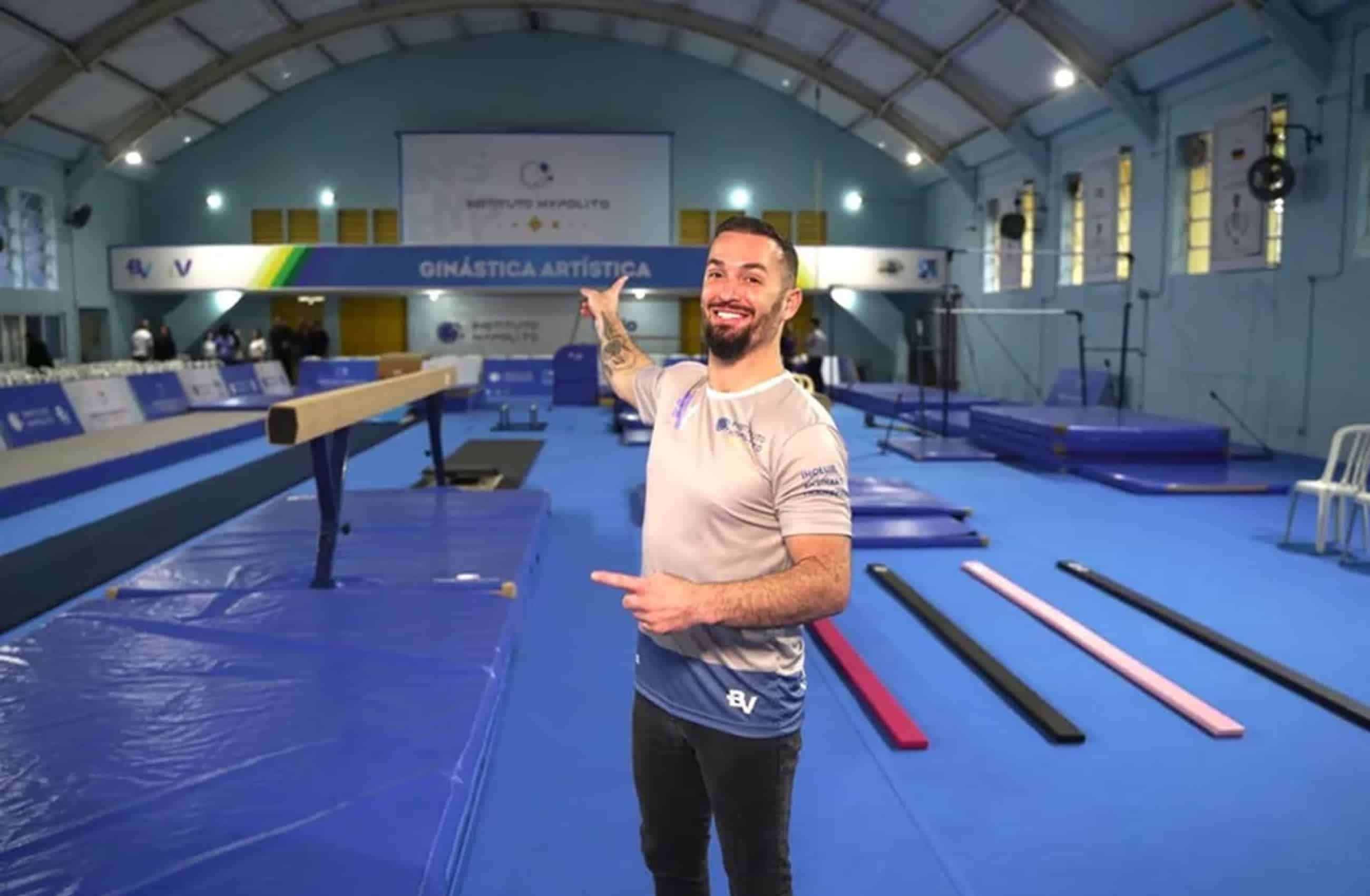 Diego Hypólito em um ginásio de ginástica artística, sorri enquanto olha para a foto e aponta para os aparelhos de ginástica.