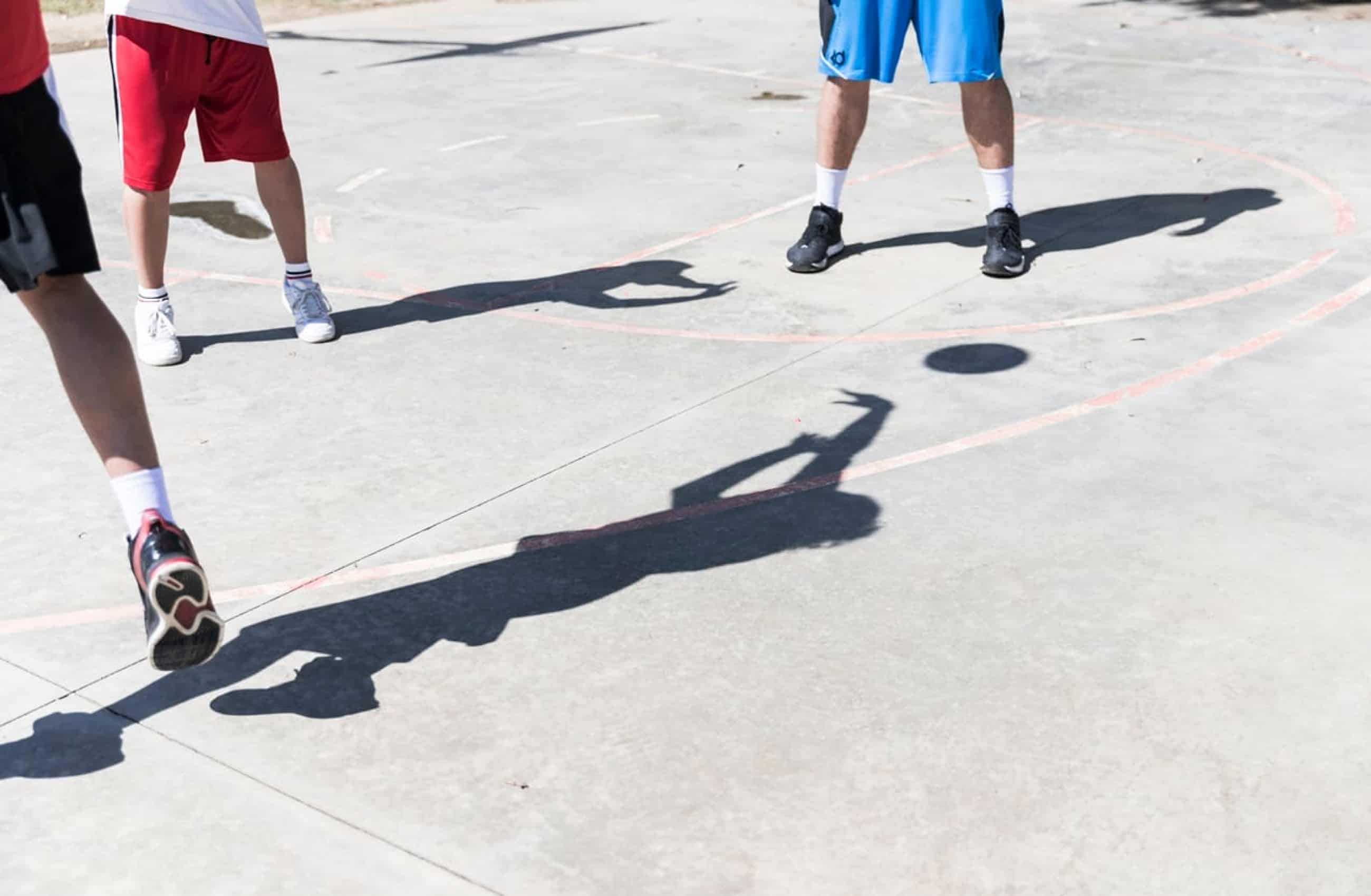 Três pessoas jogando basquete na quadra. A fotografia mostra apenas as pernas e a sombra dos jogadores.