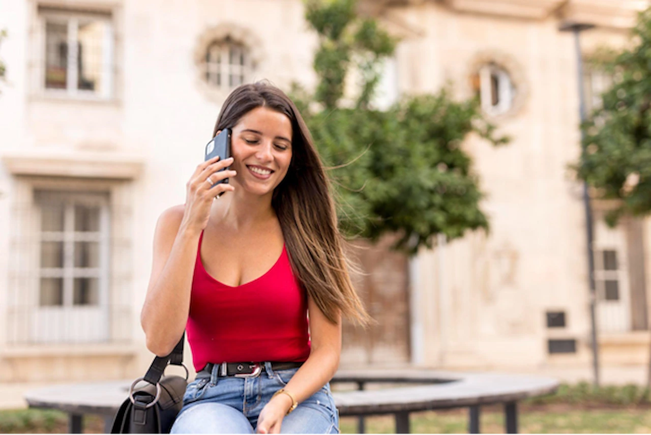 Mulher sorrindo fala ao celular em uma área externa com prédio e árvores ao fundo. 