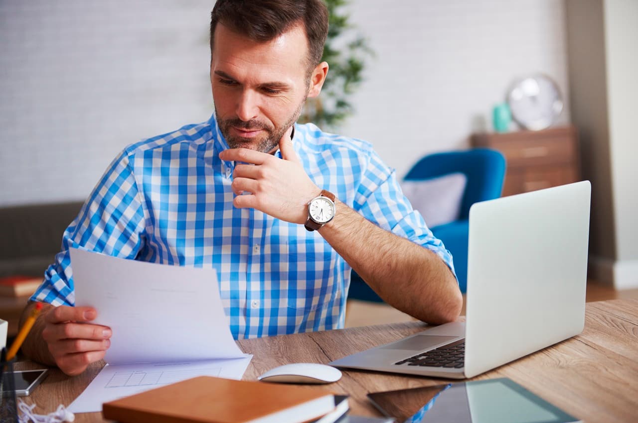 Homem pensativo, com camisa xadrez, analisando papel em seu escritório em casa, ao lado de um notebook.