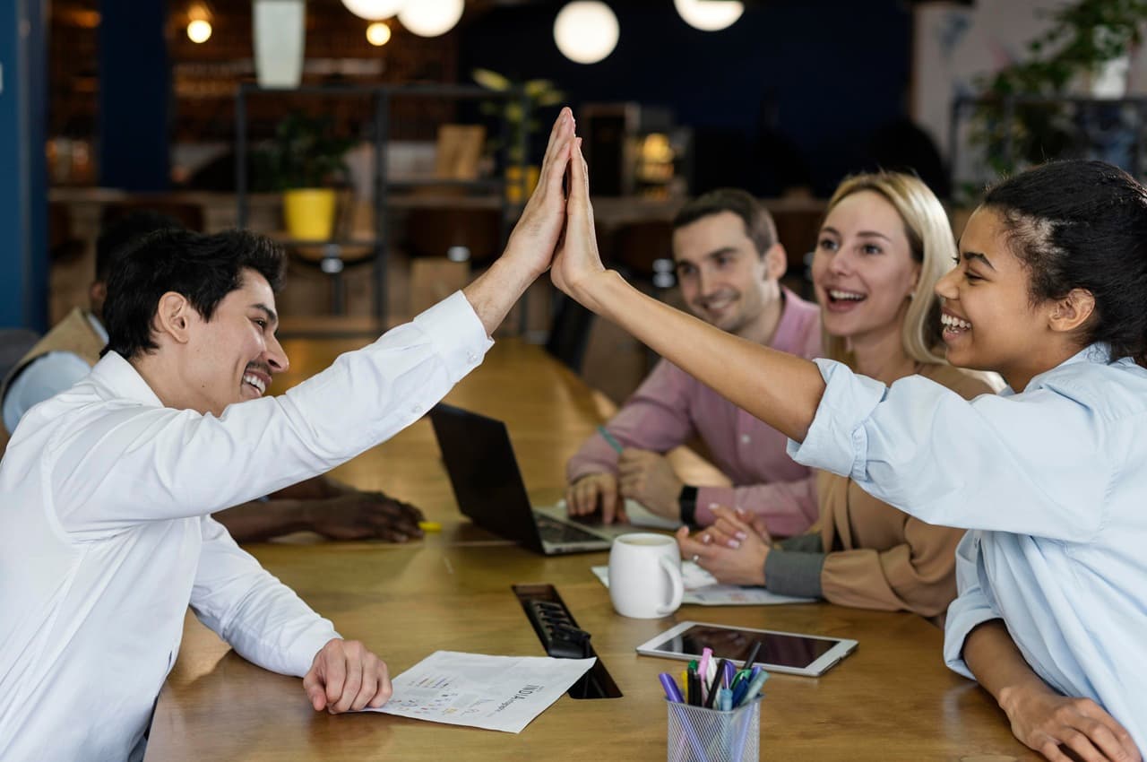 Homem e mulher sorrindo e batendo as mãos (high five) sobre uma mesa de escritório em comemoração, com colegas ao fundo.