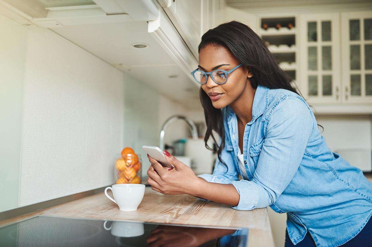 Mulher sorridente apoiada na bancada de uma cozinha, digitando no celular. Ela veste blusa jeans e calça preta.