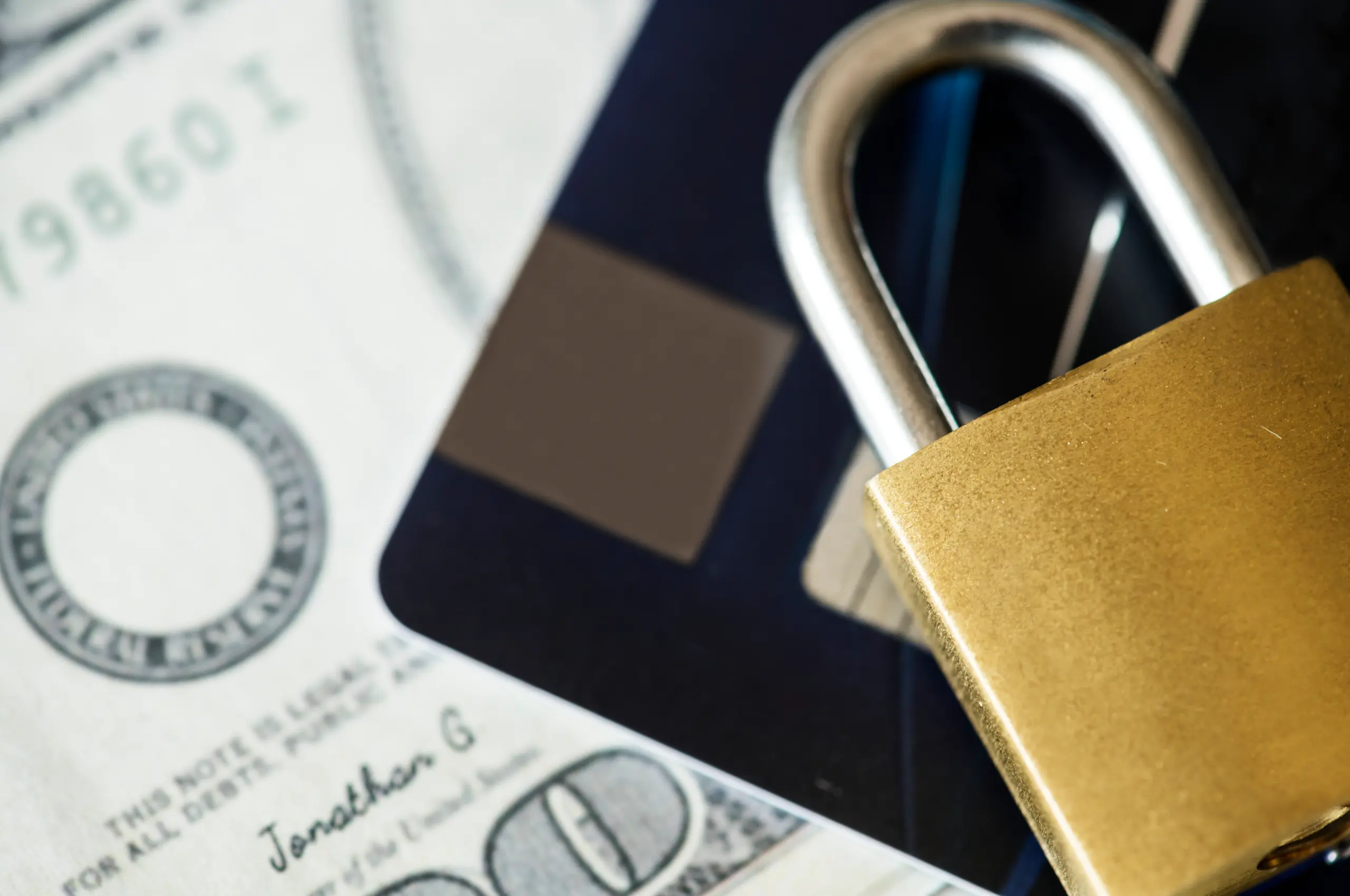 Close de uma fechadura dourada sobre notas de dólar americano, com parte de um cartão azul ao fundo, transmitindo a ideia de segurança financeira. 