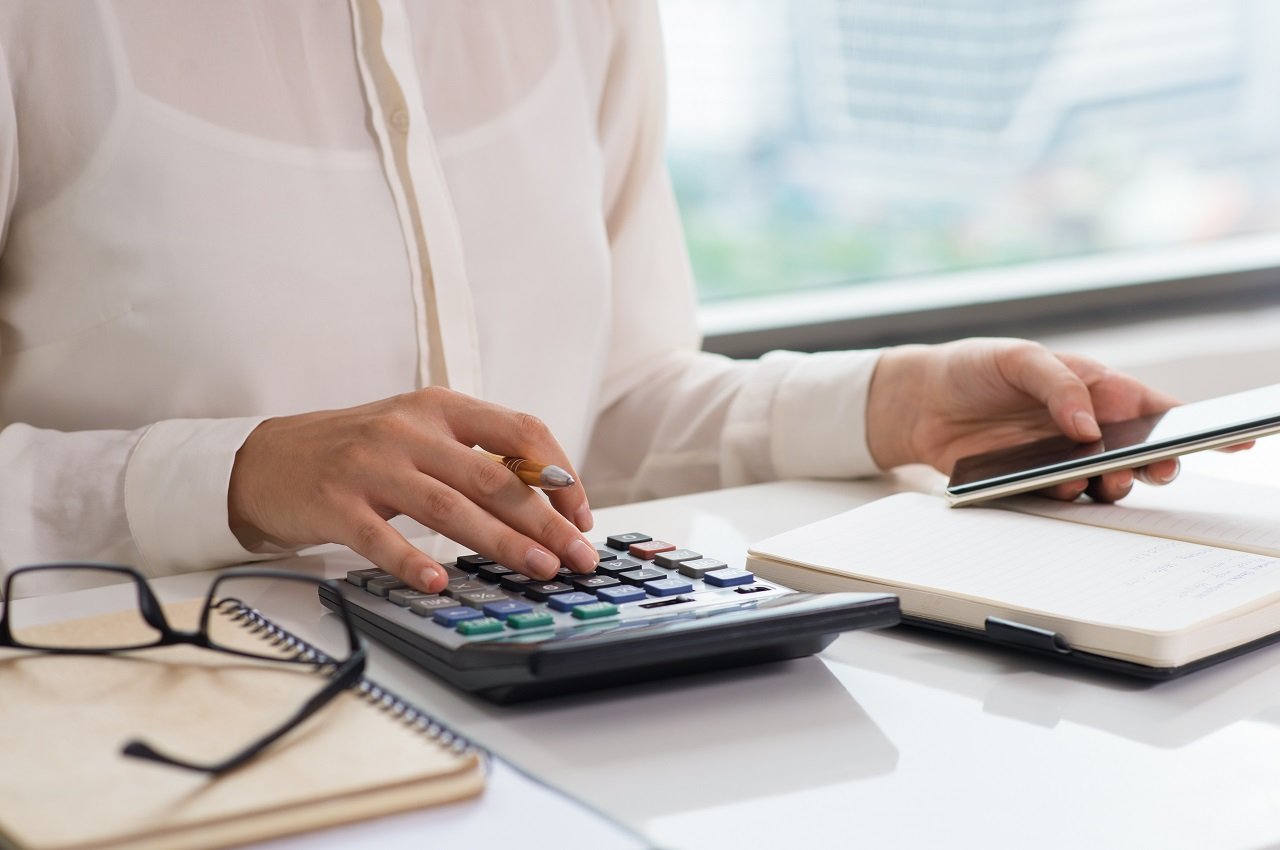 Pessoa (seu rosto não aparece na imagem) utilizando calculadora e digitando no seu computador do trabalho. A pessoa veste blusa social branca e está no escritório.