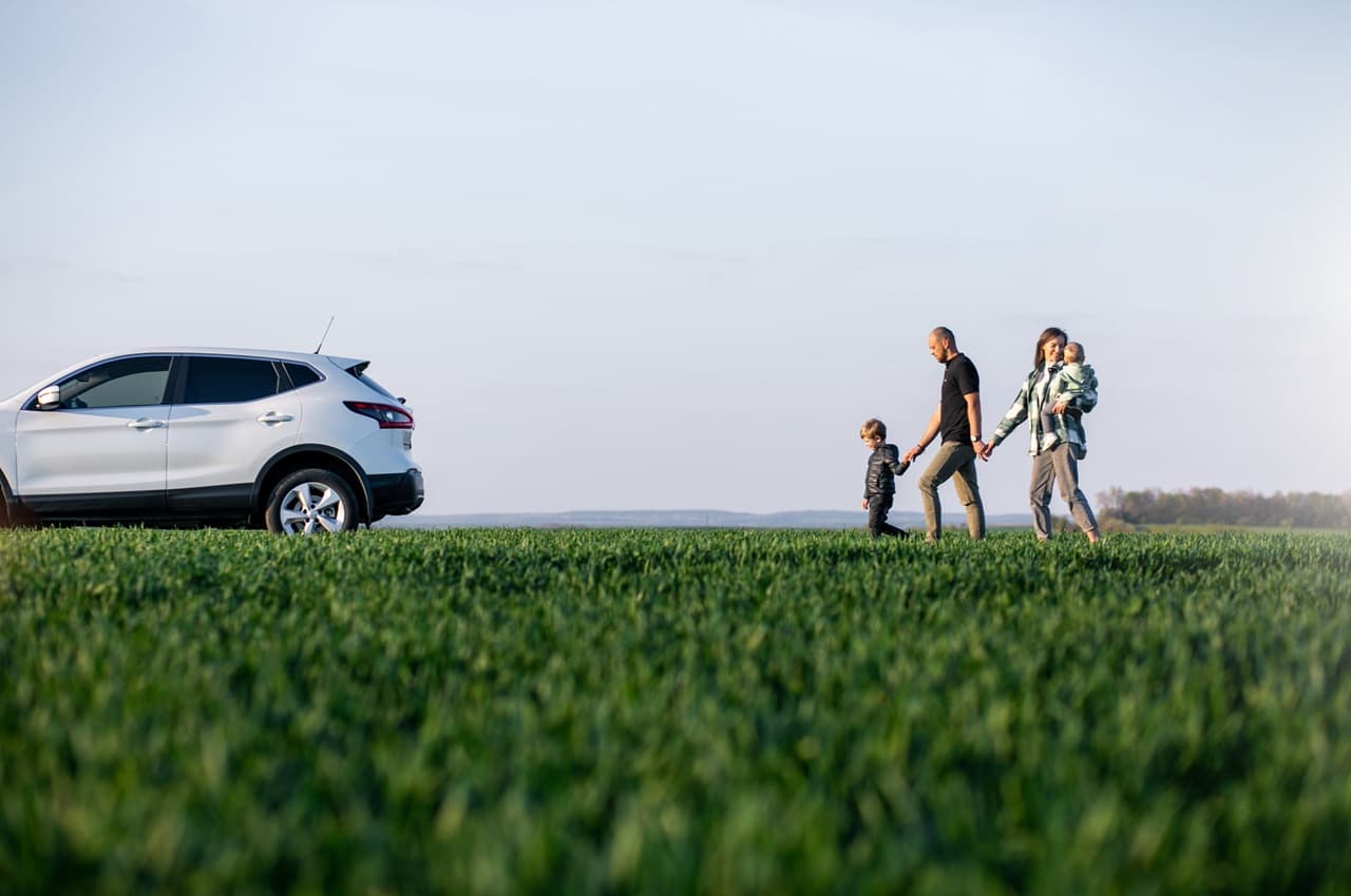 Família caminhando de mãos dadas em um campo verde aberto, com um carro branco estacionado ao fundo.
