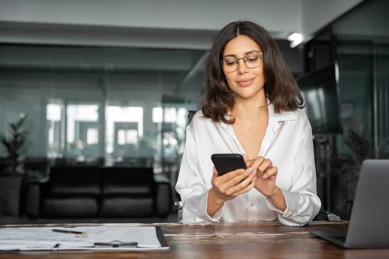 Pessoa utilizando smartphone enquanto analisa documentos sobre uma mesa com computador ao lado.