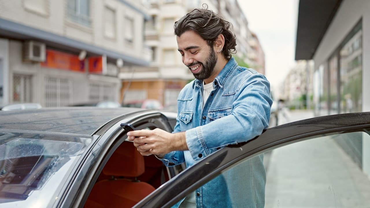Homem em pé ao lado do carro com a porta aberta, segurando o celular na mão