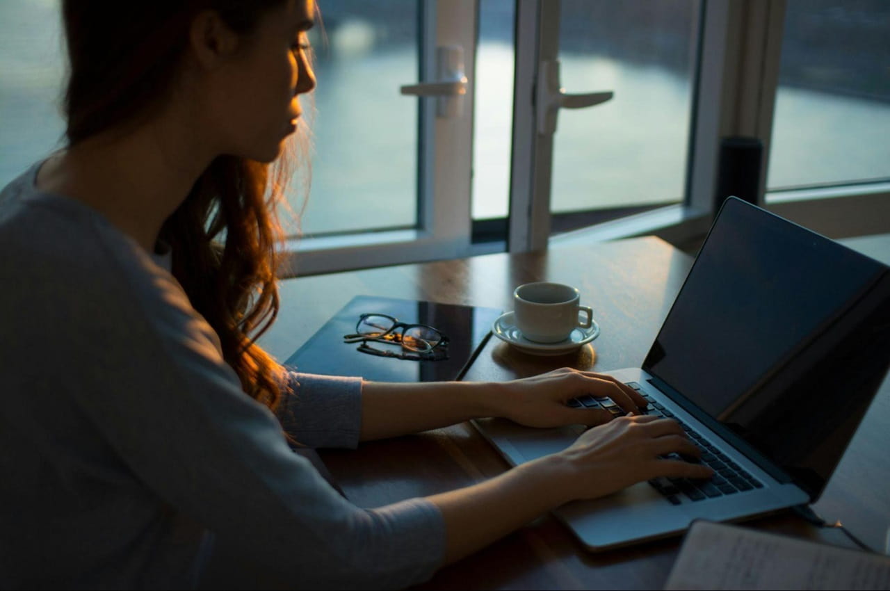 Mulher com expressão concentrada pesquisa quanto rende o FGTS no laptop em cima da mesa. A mulher tem longos cabelos castanhos e veste camisa cinza básica.