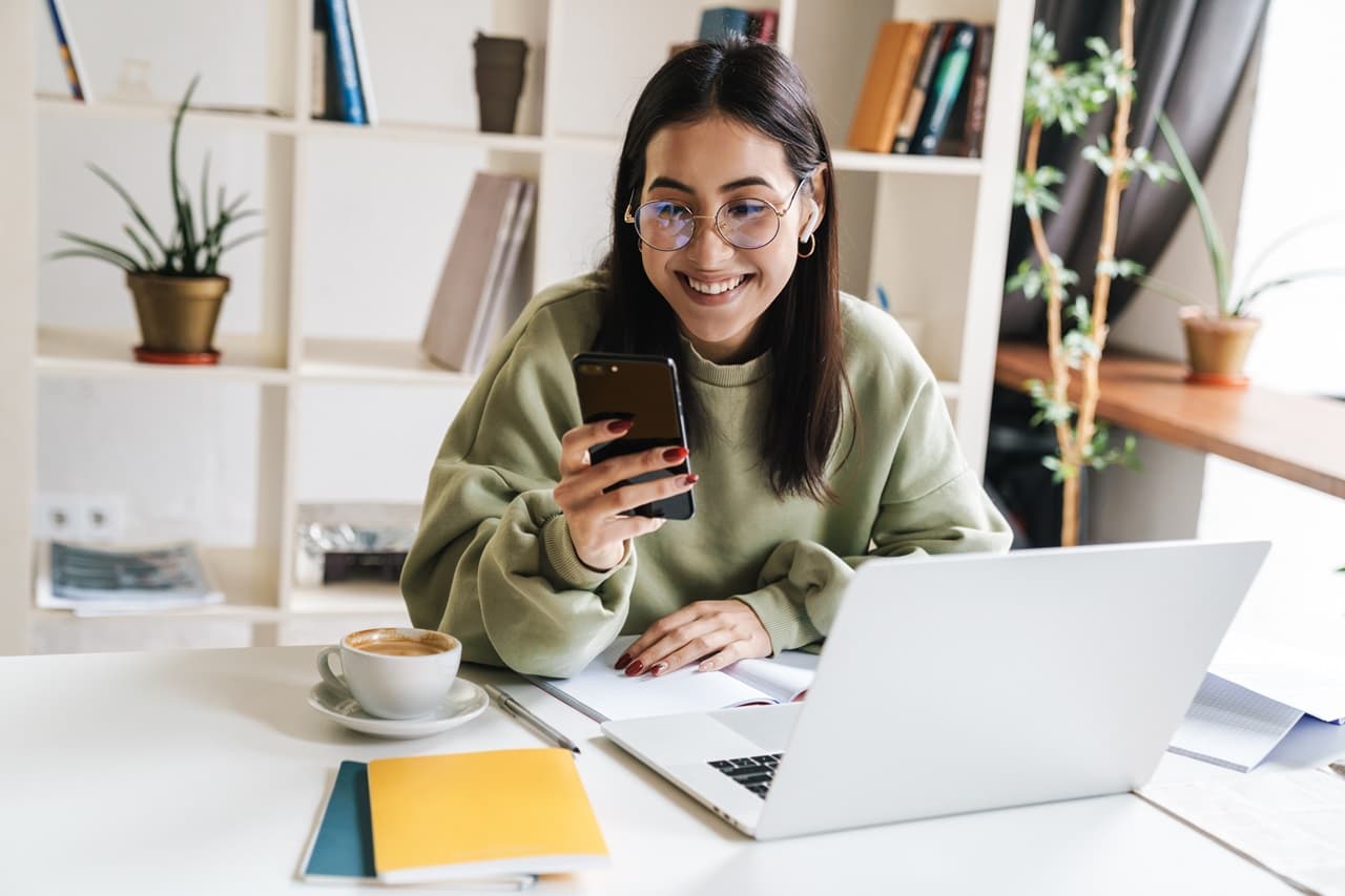 Pessoa utilizando celular e notebook em uma mesa durante consulta online de informações financeiras.