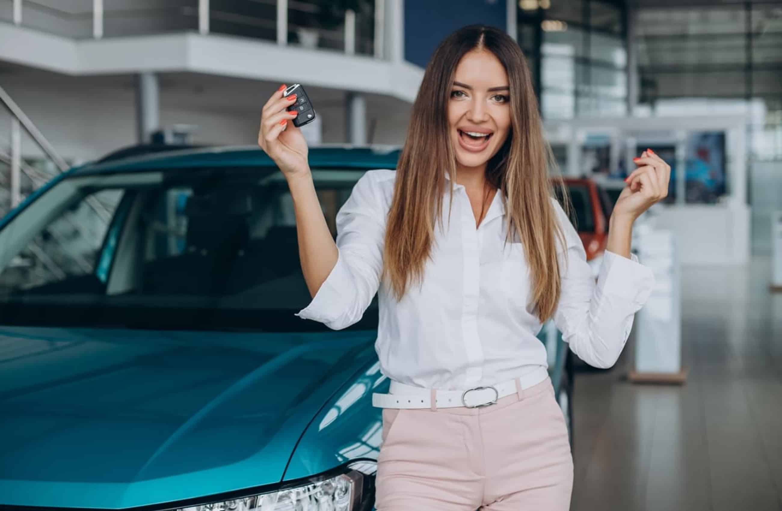Mulher de cabelos castanhos, camisa social branca e calça rosa claro está sorrindo com a boca aberta enquanto dá risada e olha para a câmera. Ao fundo temos a imagem de um carro azul esverdeado. A imagem representa o Financiamento de Veículos BV.