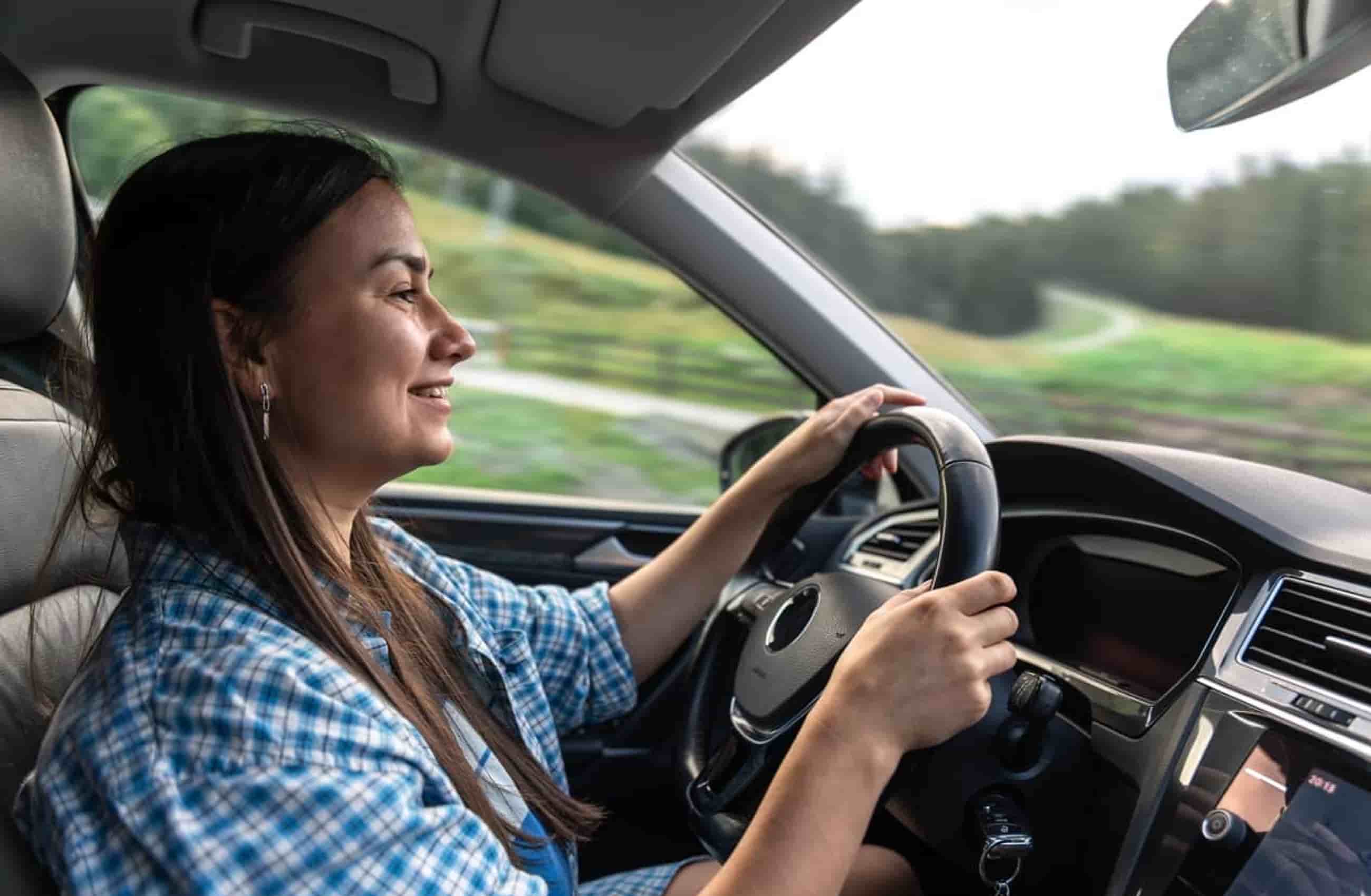 Mulher de cabelos castanhos e blusa xadrez azul está dirigindo um carro enquanto sorri. A imagem mostra seu perfil, e a foto parece ter sida tirada do banco do passageiro. A imagem representa quanto custa manter um carro.
