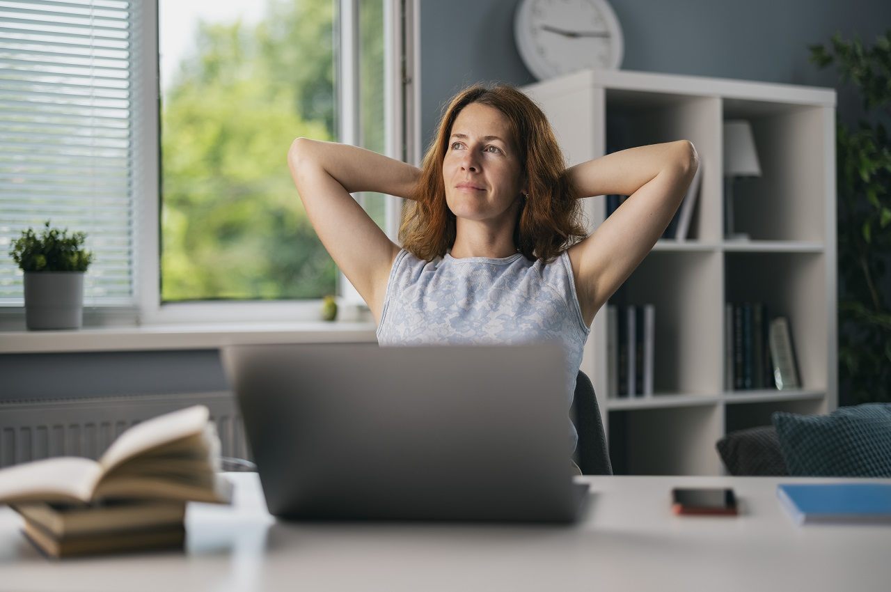 Mulher sorridente sentada no escritório e apoiando os braços atrás da cabeça, enquanto inclina sua cadeira. Ela olha para a tela de um computador.