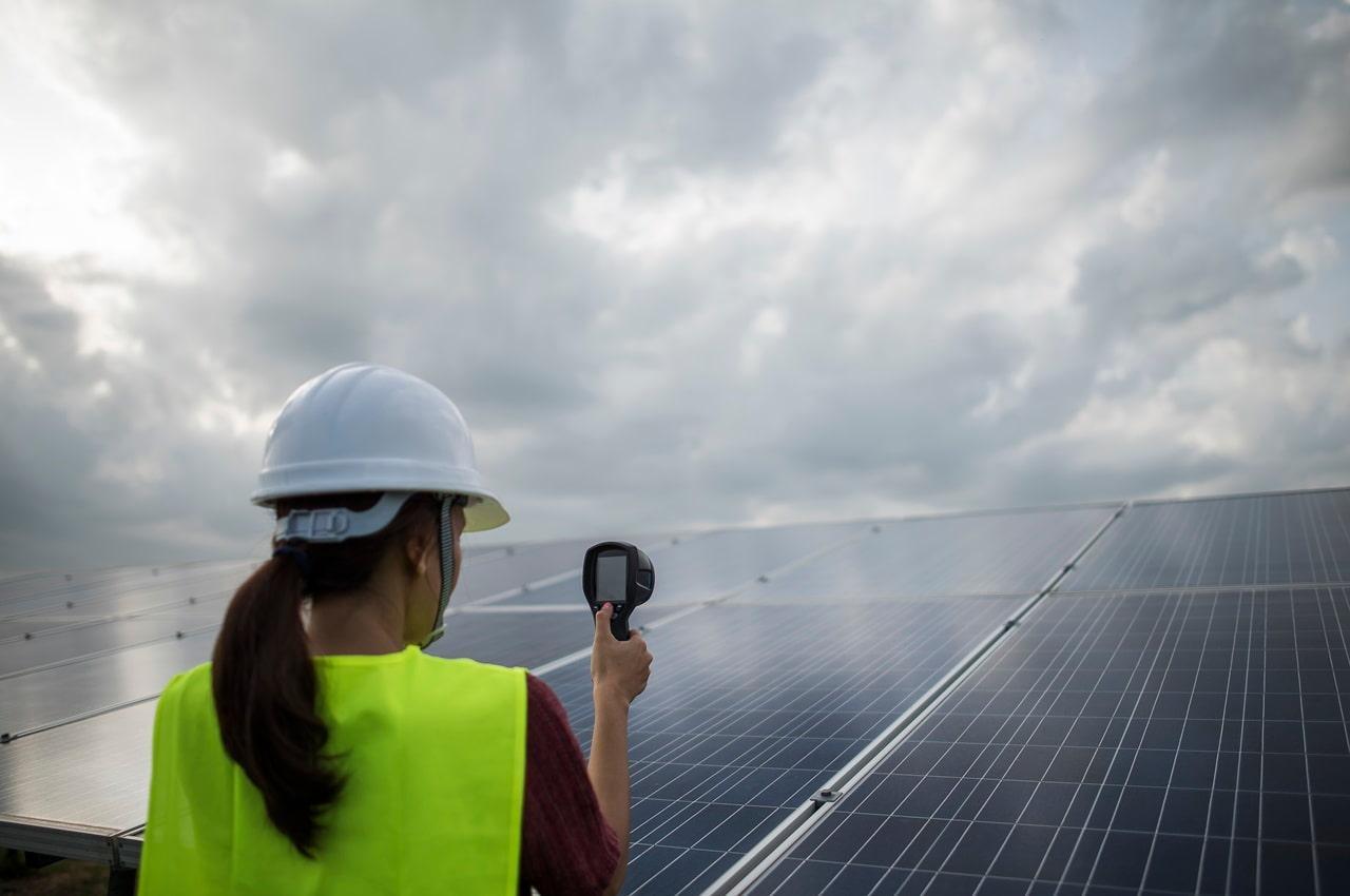 Mulher inspecionando painéis solares com um aparelho de medição, sob um céu claro. Ela está usando um colete verde e um capacete de segurança.
