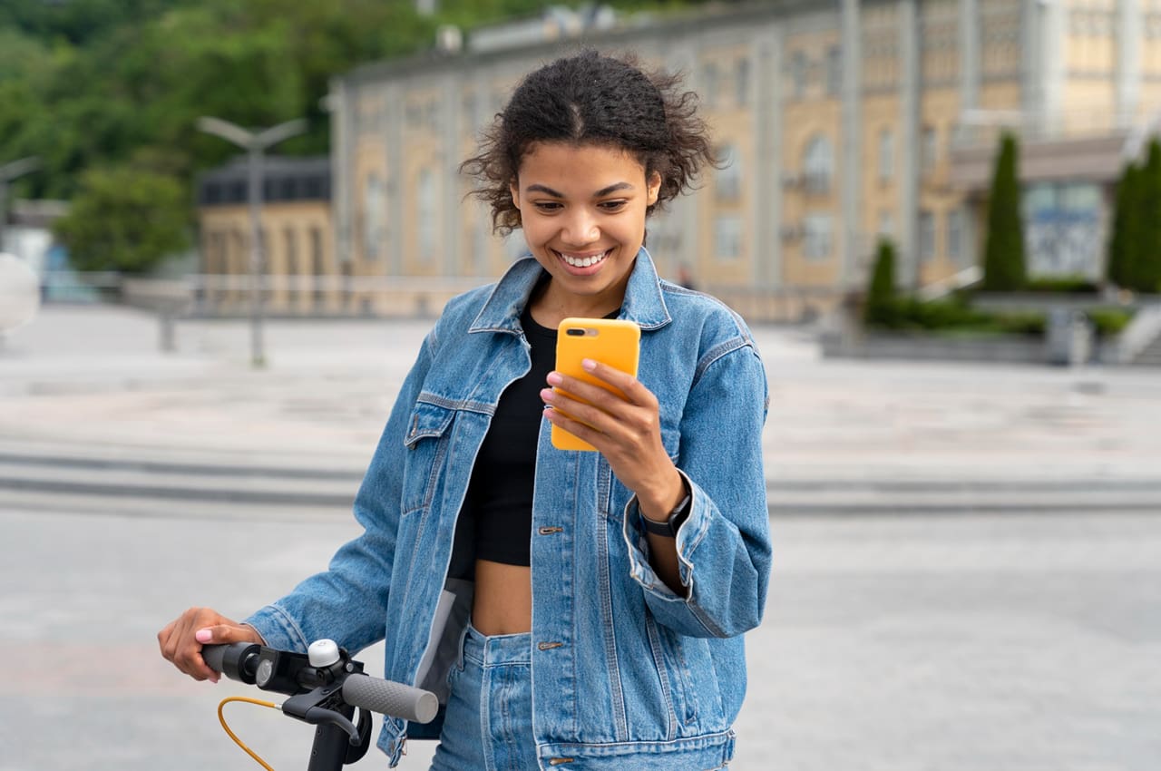 Mulher negra sorridente em um patinete elétrico, simbolizando a mobilidade sustentável. Ela está olhando para o seu telefone celular.