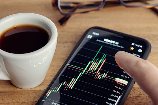 Xícara de café ao lado de um smartphone exibindo gráficos financeiros, com uma mão apontando para os dados na tela. Óculos estão visíveis ao fundo. 
