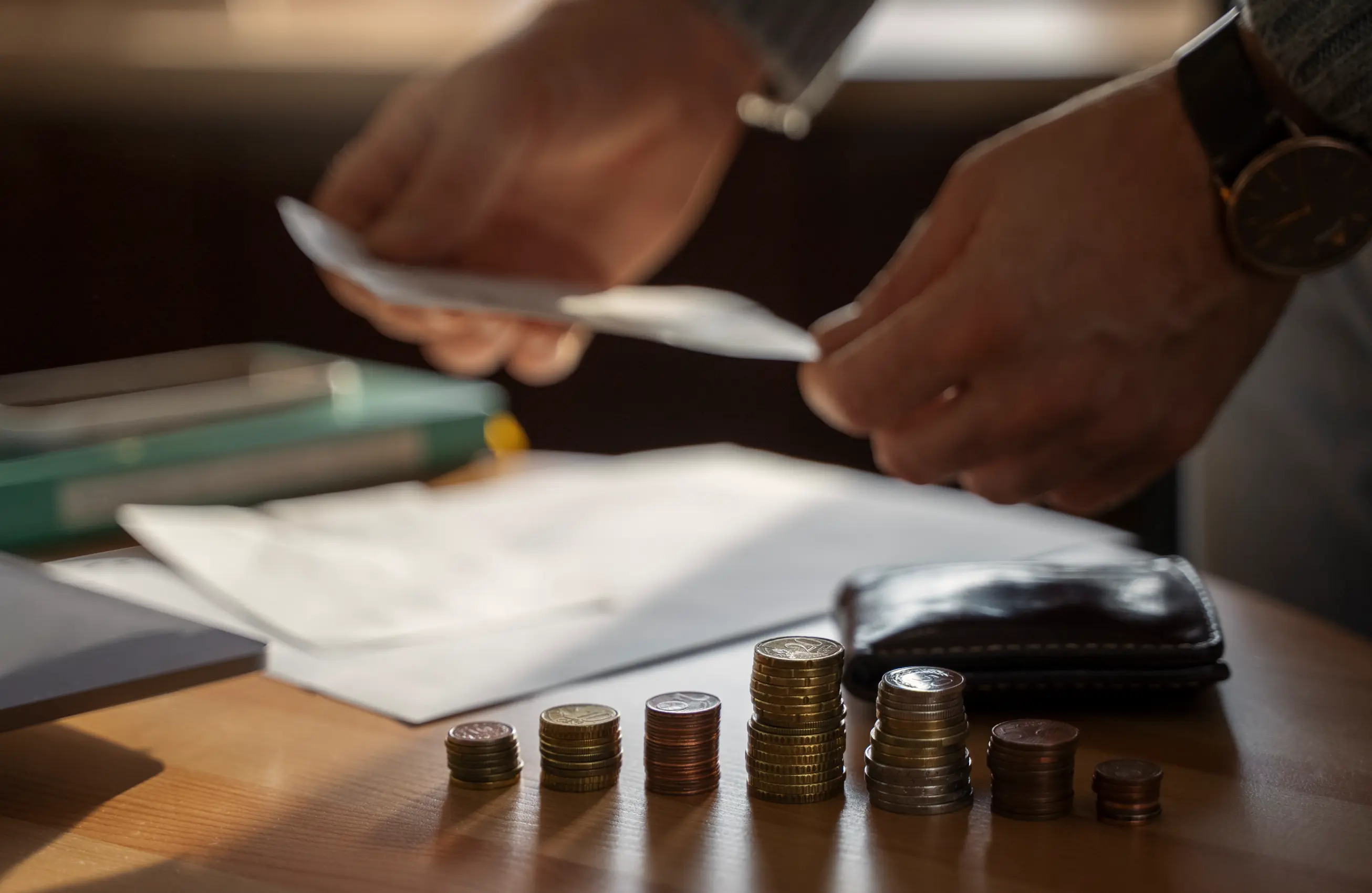 Mãos de uma pessoa segurando papéis sobre uma mesa de madeira, onde há pilhas organizadas de moedas, uma carteira de couro e documentos, sugerindo o conceito de organização financeira ou planejamento de despesas. 