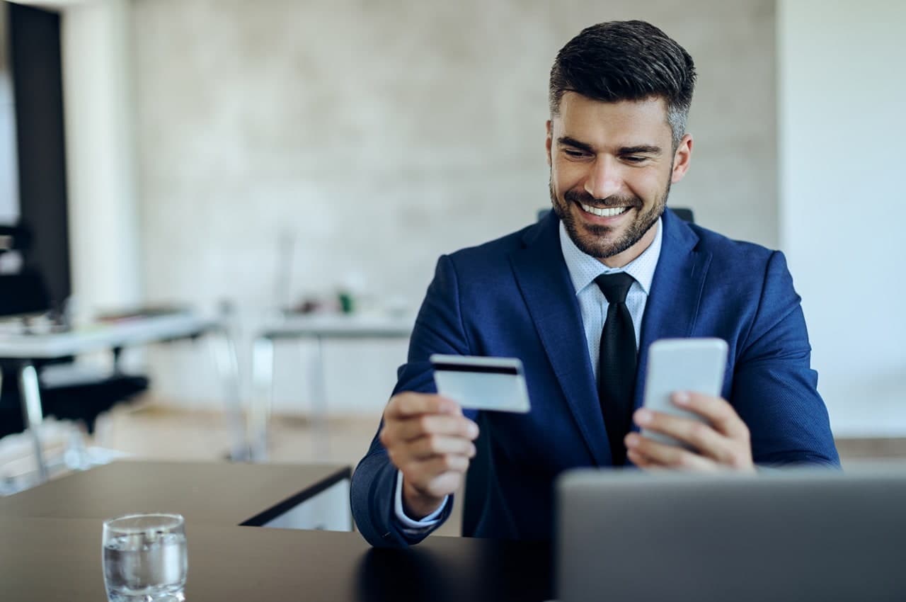 Homem de terno azul-escuro segurando um cartão de crédito e um celular, enquanto está sentado em uma mesa de escritório.