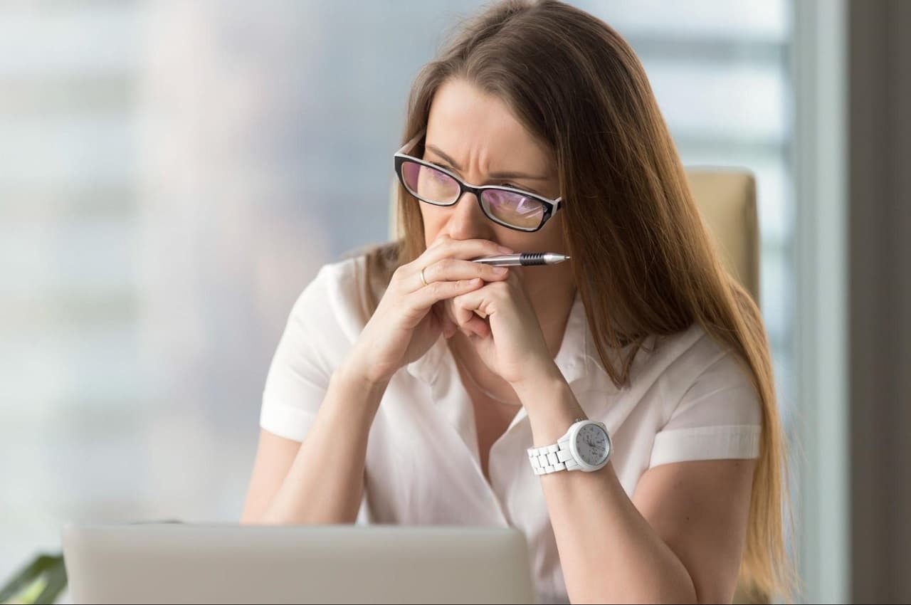 Mulher com expressão concentrada, juntando as sobrancelhas e com as mãos fechadas sob a boca. Ela olha para a tela de um computador.