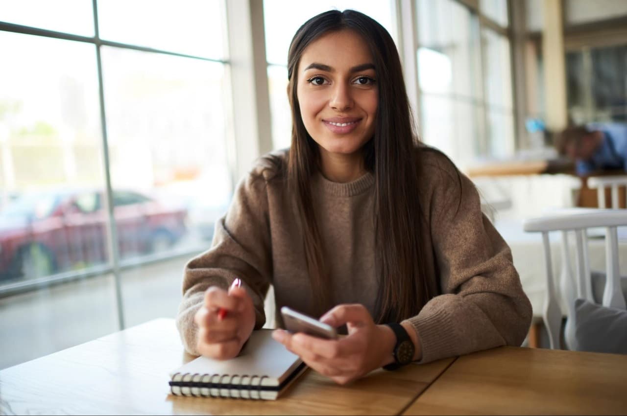 Mulher sorridente de cabelos e olhos castanhos, utilizando celular para saque-aniversário. Ela veste moletom marrom e está sentada em uma cafeteria.