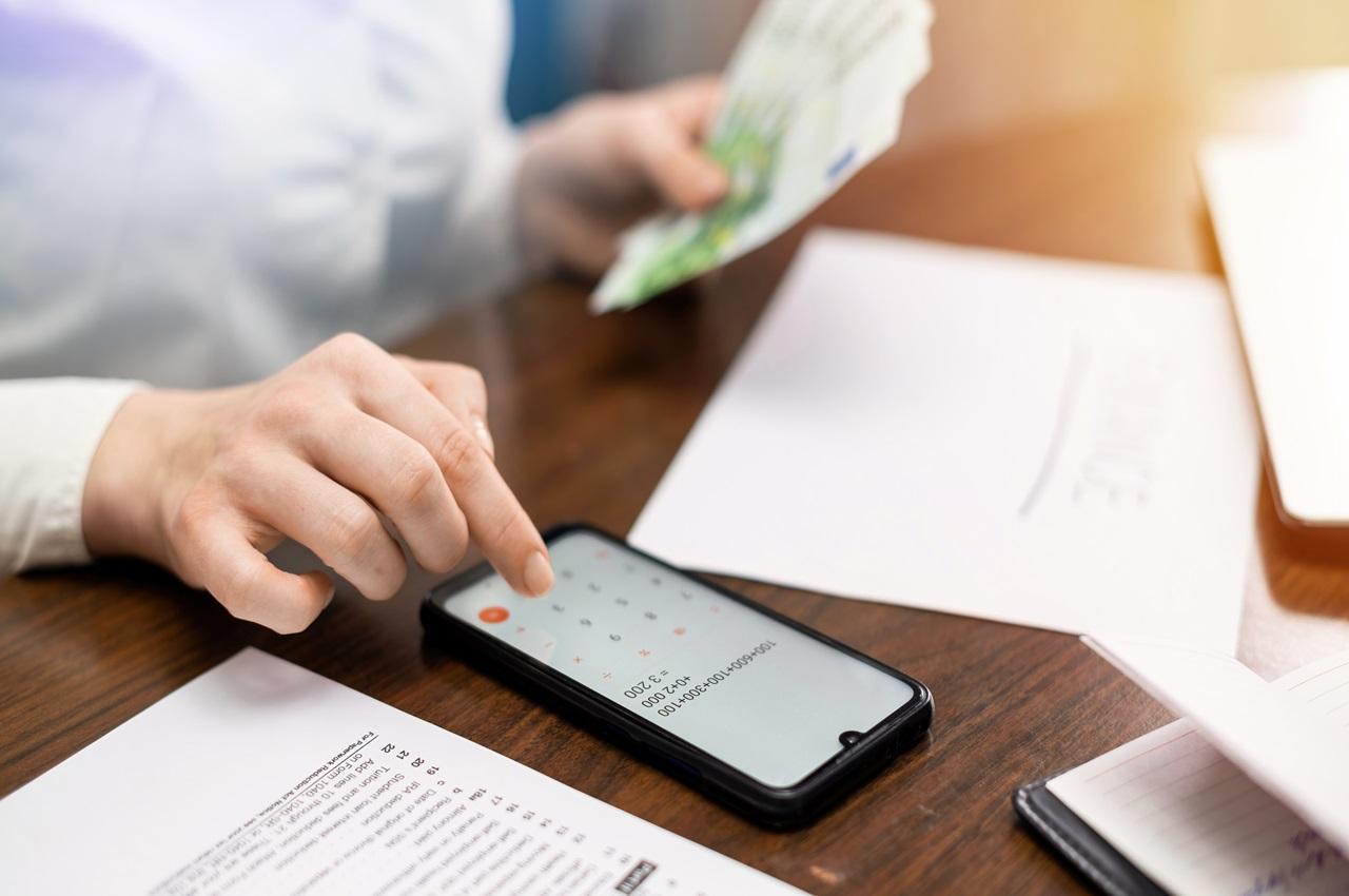 Pessoa (o rosto não aparece na imagem) calculando o saque imediato FGTS na calculadora do celular e segurando vários papéis na mão.