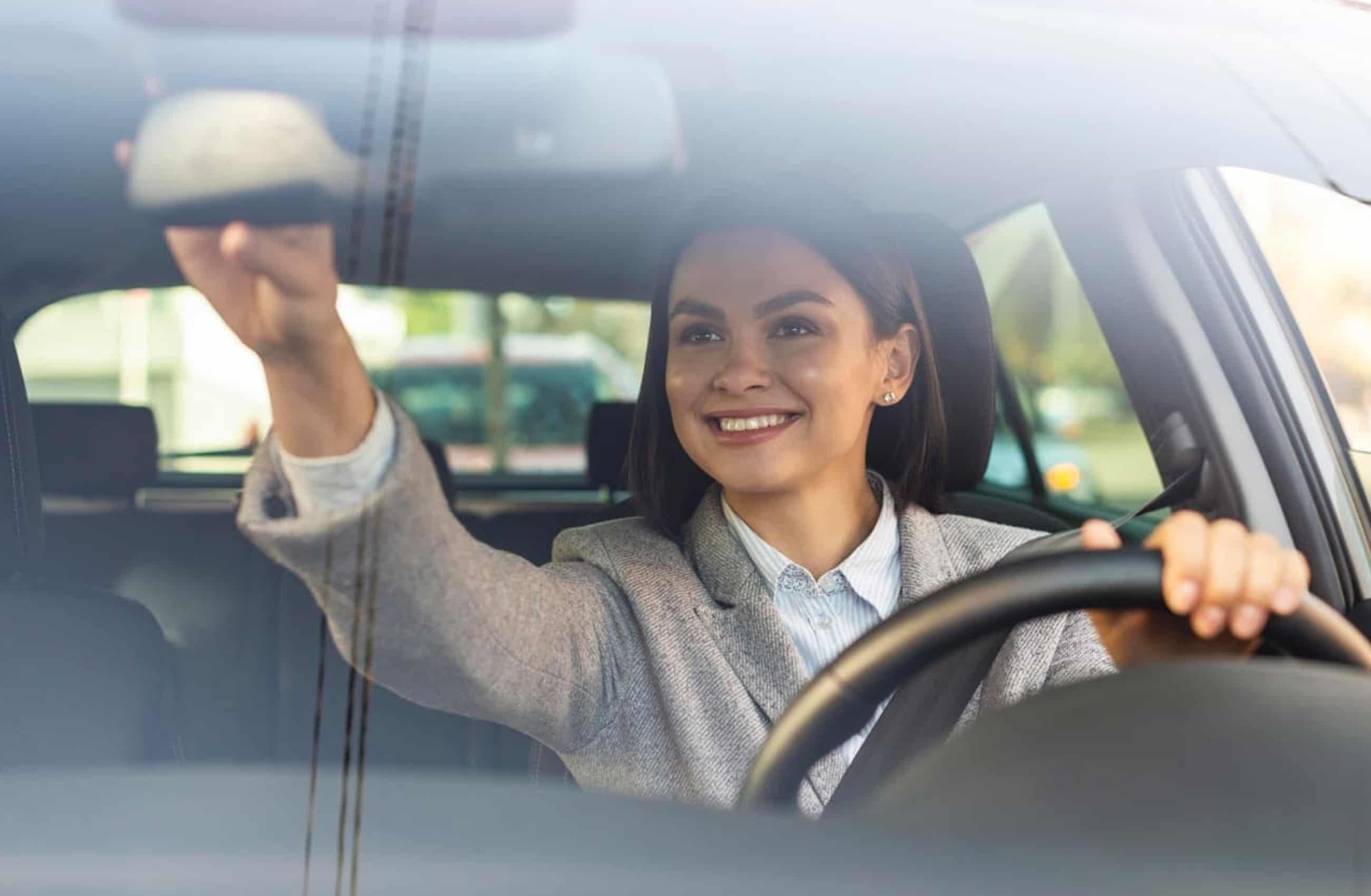 Mulher branca sentada no banco do motorista dentro de um carro. Ela ajusta o retrovisor interno enquanto sorri para o reflexo.