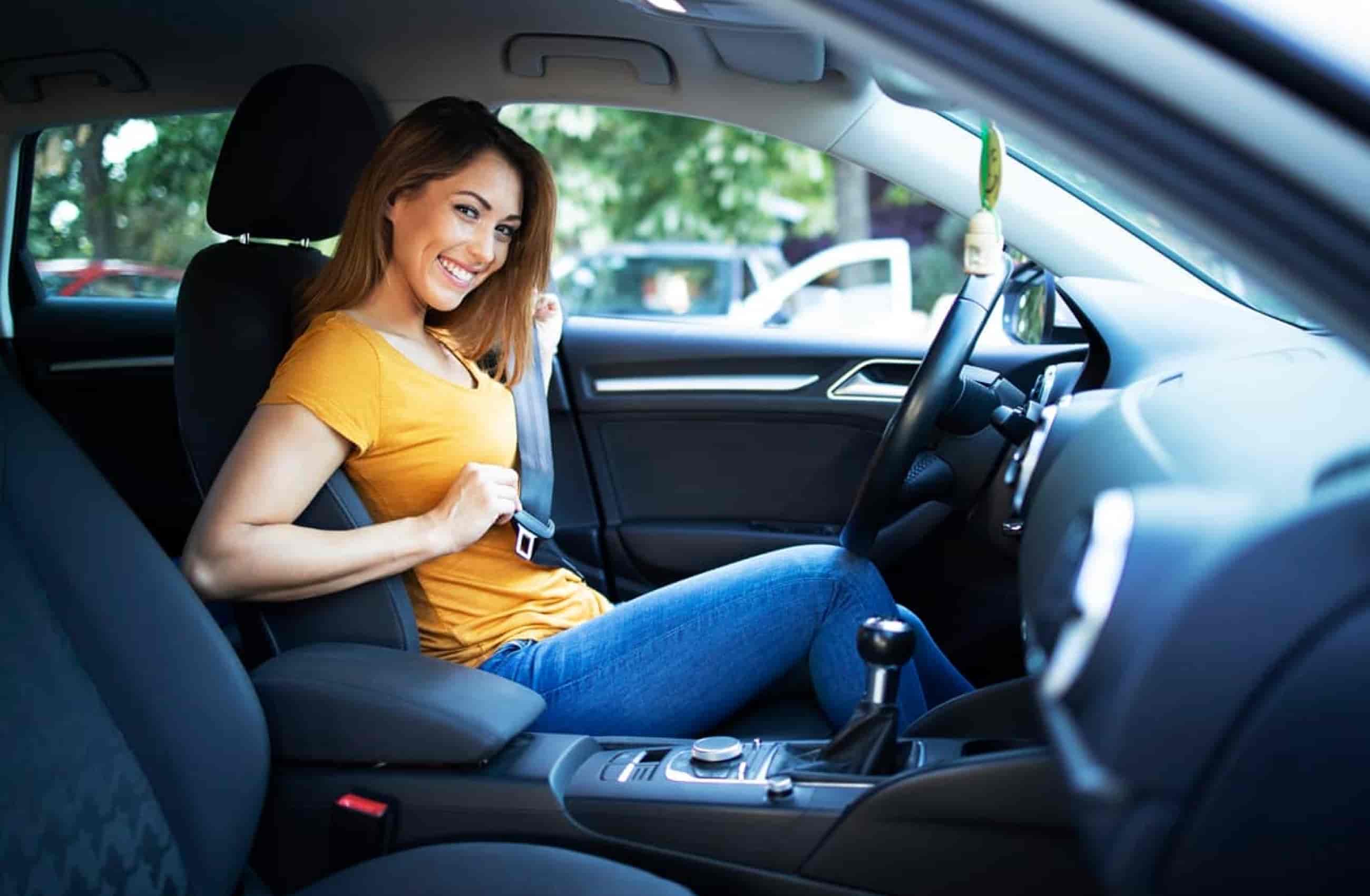 Mulher branca de blusa amarelo e calça jeans está sorrindo sentada no banco do motorista, enquanto coloca um cinto de segurança dentro de um carro.