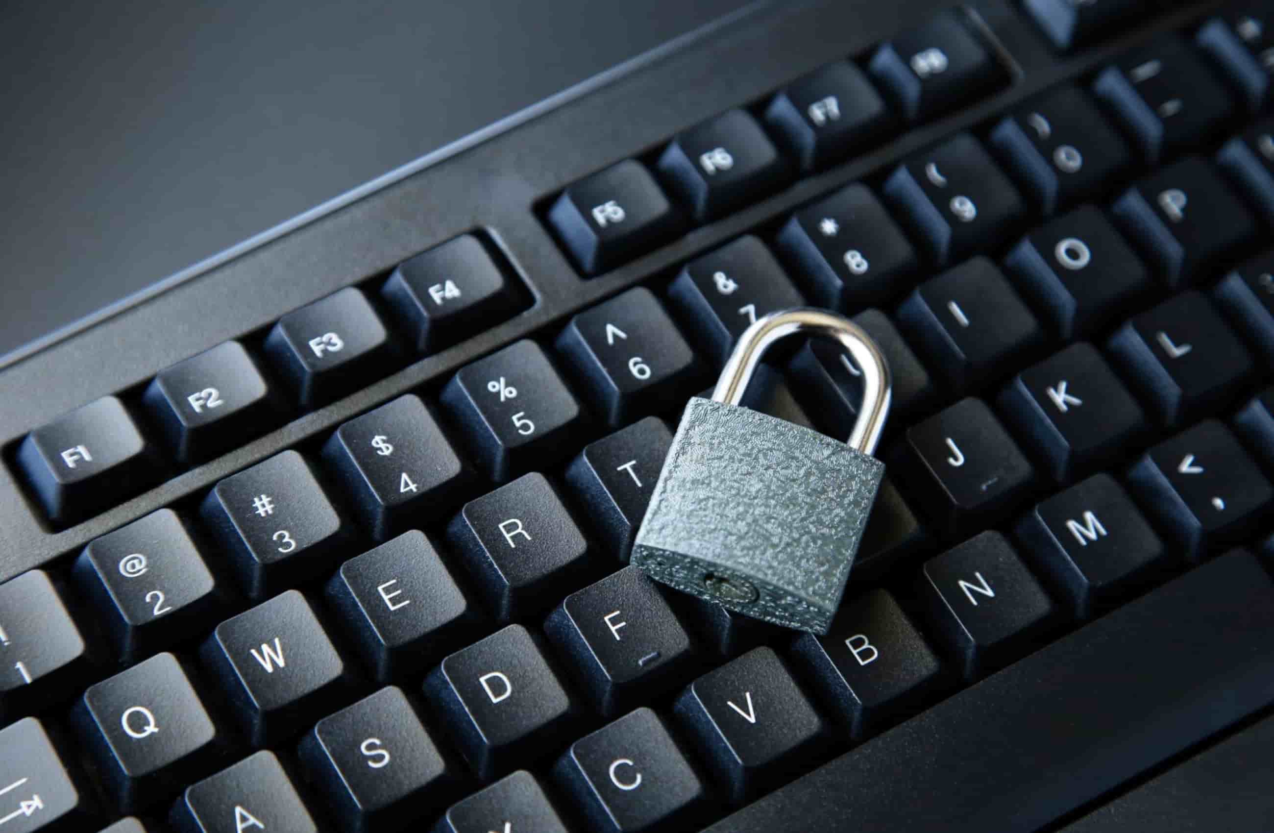 Um cadeado de metal cinza com haste cromada está posicionado em ângulo sobre as teclas pretas de um teclado de computador, simbolizando a importância da senha forte. 