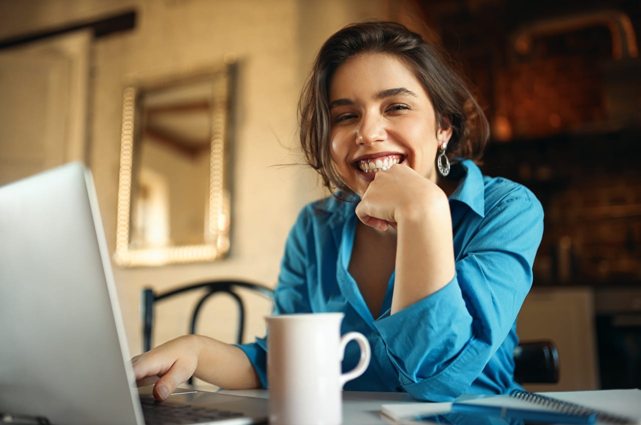 Mulher sorridente sentada à mesa com um notebook, uma caneca e um caderno. Ela está pesquisando sobre o Simulador de Empréstimo com Garantia de Veículo.