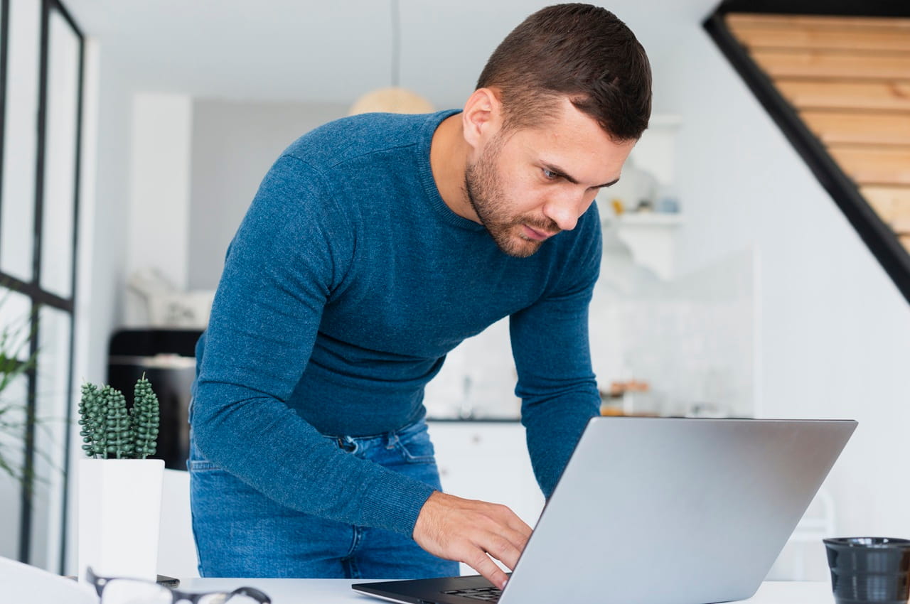 Homem de blusa azul em pé em um ambiente moderno e bem iluminado. Ele está inclinado sobre uma mesa com um notebook.