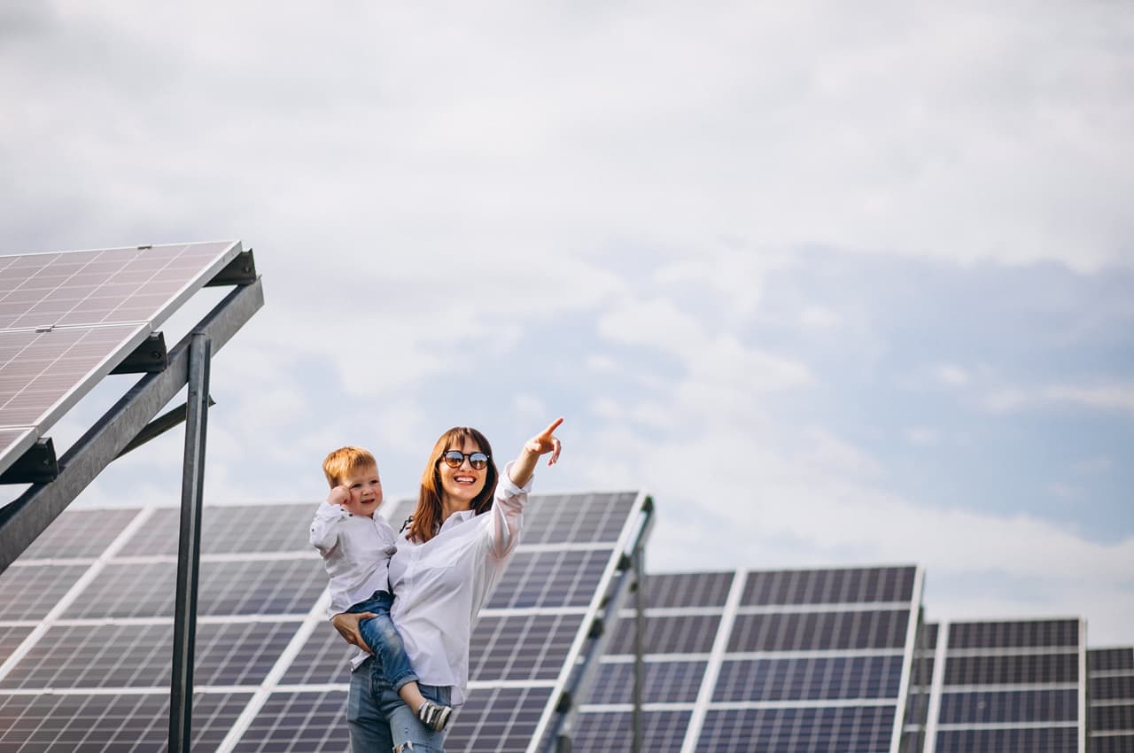 Mulher de óculos escuros com uma criança no colo, apontando para o céu em frente a painéis solares instalados.