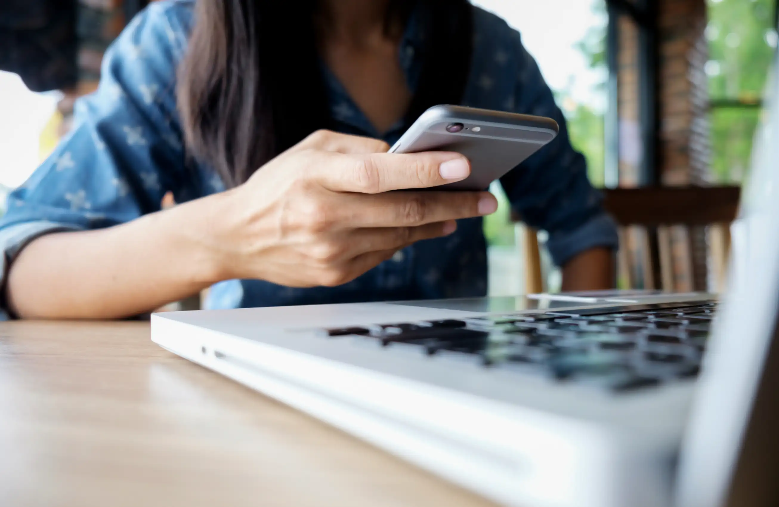 Pessoa segurando um smartphone com a mão direita enquanto usa um laptop sobre a mesa; foco nas mãos e nos dispositivos, ambiente tipo café ao fundo.&nbsp;