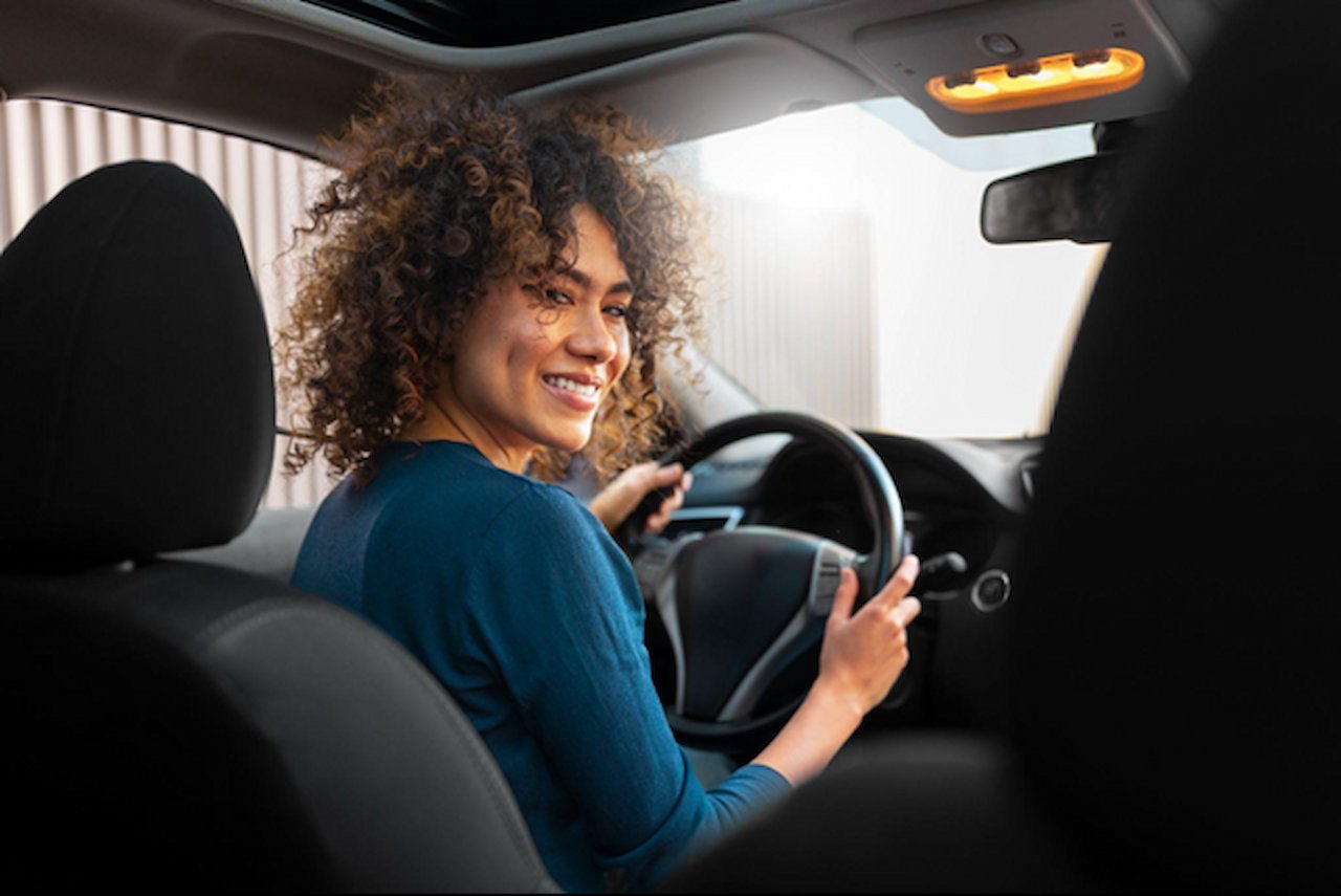 Pessoa sorrindo ao volante, olhando pelo ombro enquanto dirige um carro, vista do banco traseiro com volante e painel ao fundo.
