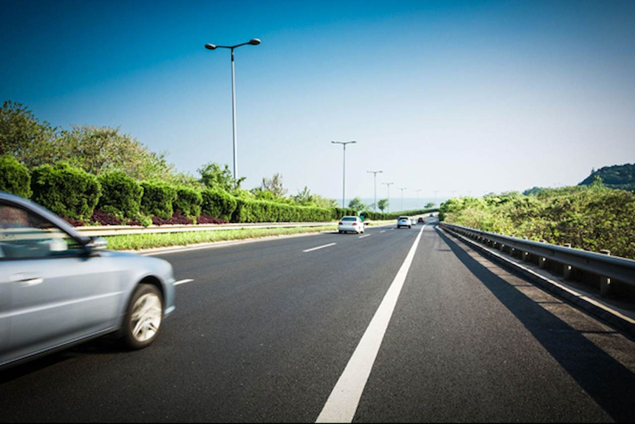 Rodovia de pista dupla em dia ensolarado, com alguns carros trafegando e barreira metálica à direita; vegetação e postes ao longo da via.