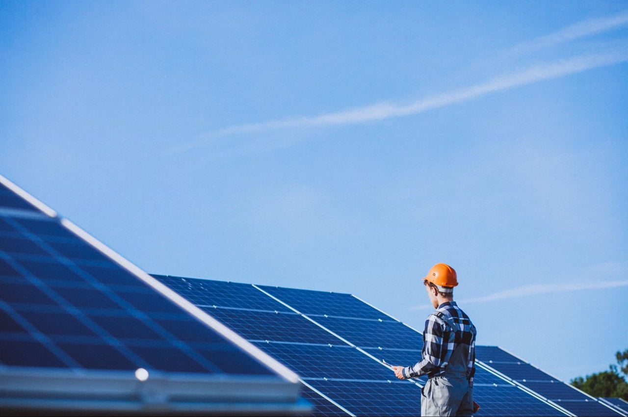 Técnico, de chapéu e uniforme, inspecionando painéis solares no telhado de uma instalação ao ar livre.