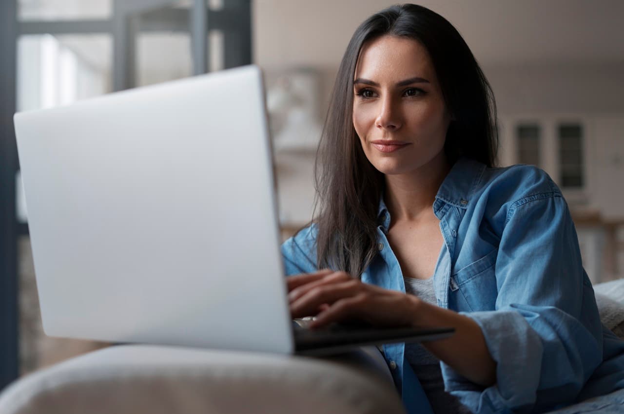 Mulher de cabelos escuros e camisa jeans usando um laptop. Ela está sentada confortavelmente em um sofá e pesquisando sobre tarifa de cadastro em empréstimo.