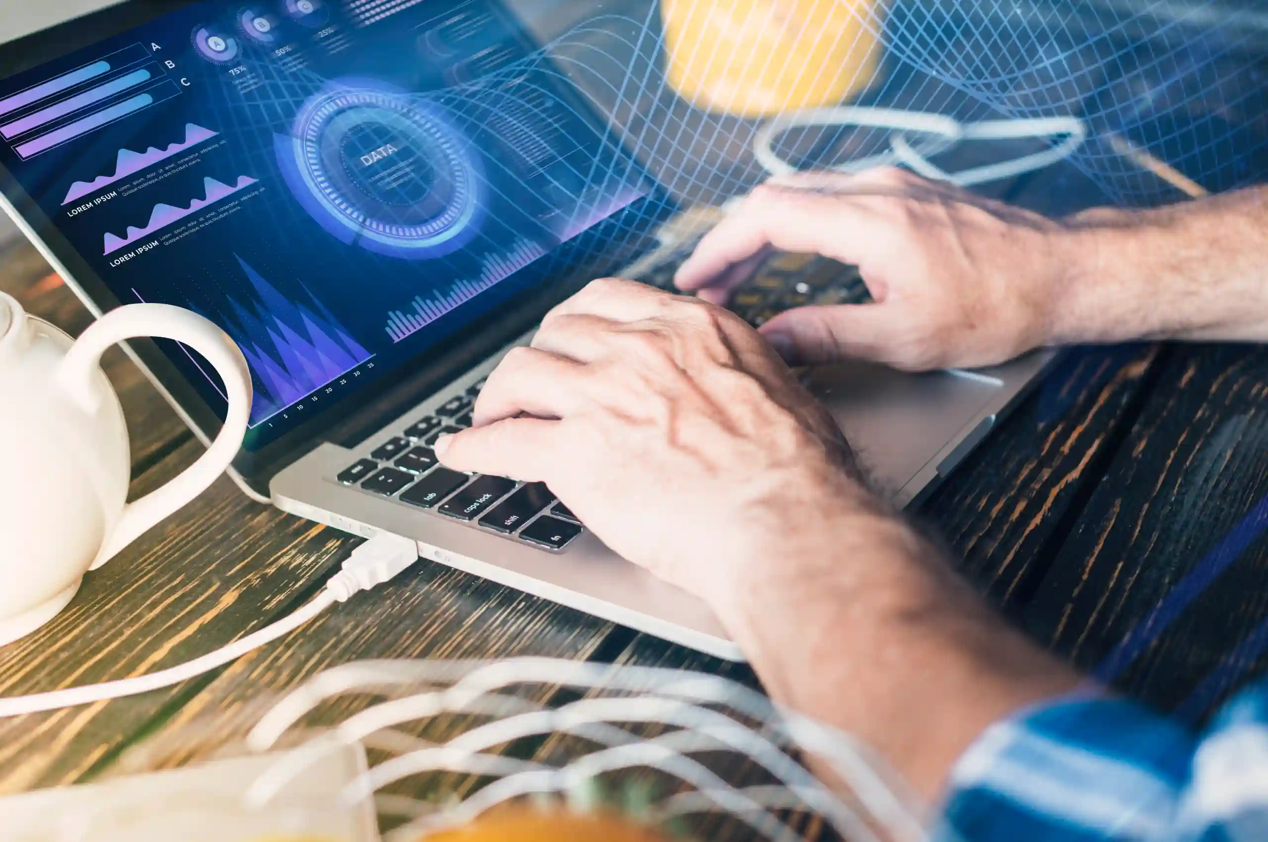 Homem digitando em um notebook sobre uma mesa de madeira, com gráficos e dados digitais exibidos na tela do computador. Ao lado, há uma xícara branca e fios conectados ao notebook. A imagem transmite um ambiente de análise de dados ou trabalho com tecnologia.