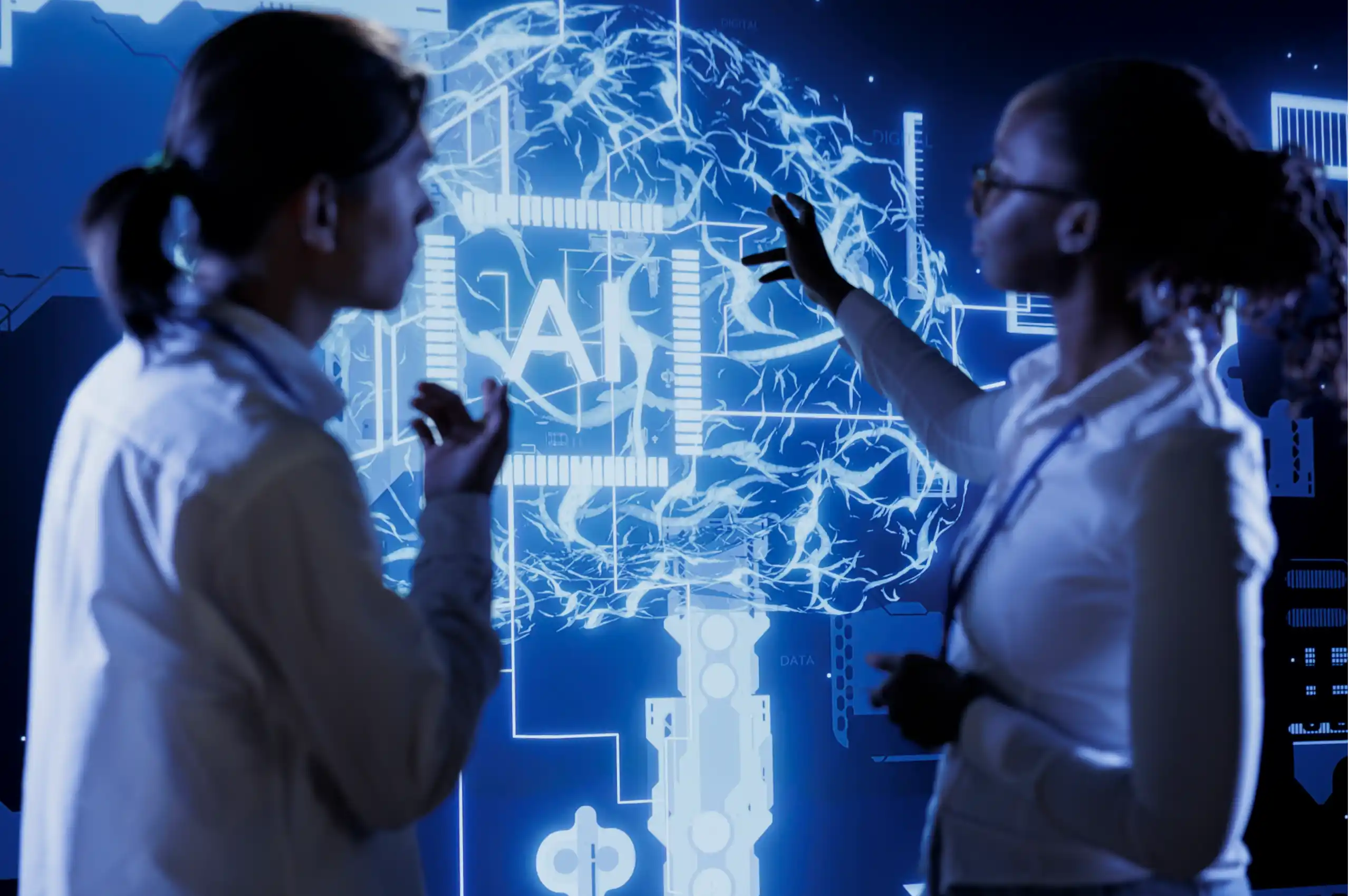 Duas pessoas em pé, de perfil, conversando e apontando para uma grande tela digital azul que exibe a imagem de um cérebro estilizado com circuitos e a sigla "AI" (inteligência artificial) no centro, sugerindo um ambiente de tecnologia ou inovação.