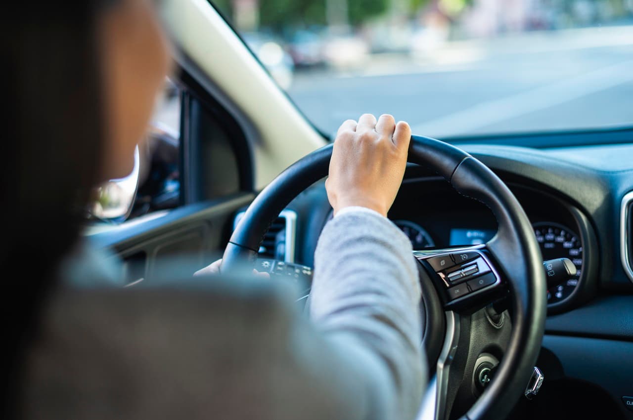 Mão de uma pessoa ao volante, dirigindo um veículo quitado pela cidade.