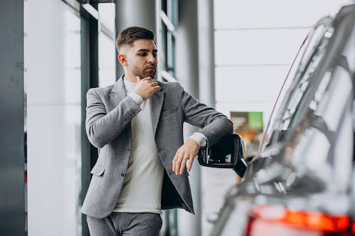Homem com um braço apoiado no retrovisor de um carro e outra mão no queixo.