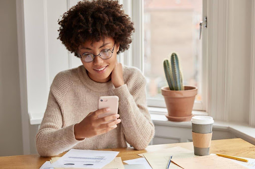 Uma mulher sentada sobre uma mesa com diversos papéis enquanto utiliza um smartphone.