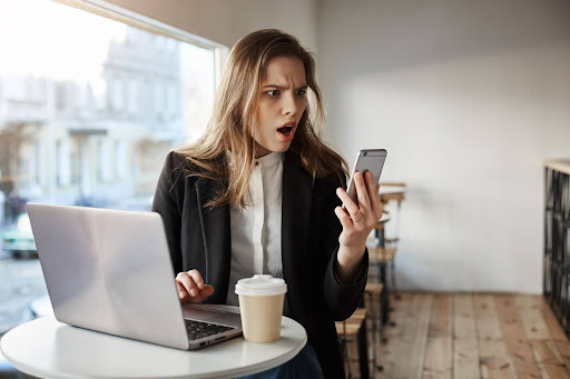 Uma mulher sentada em uma cafeteria enquanto encara um smartphone chocada. 