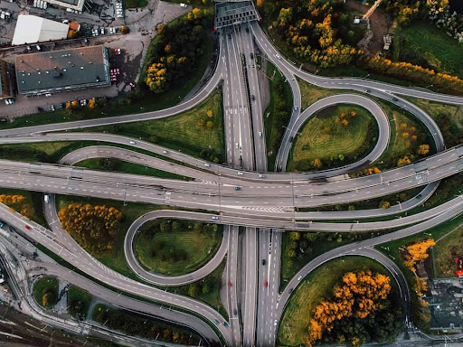 Vista aérea de um entroncamento rodoviário com várias alças e viadutos entre áreas verdes; carros em circulação