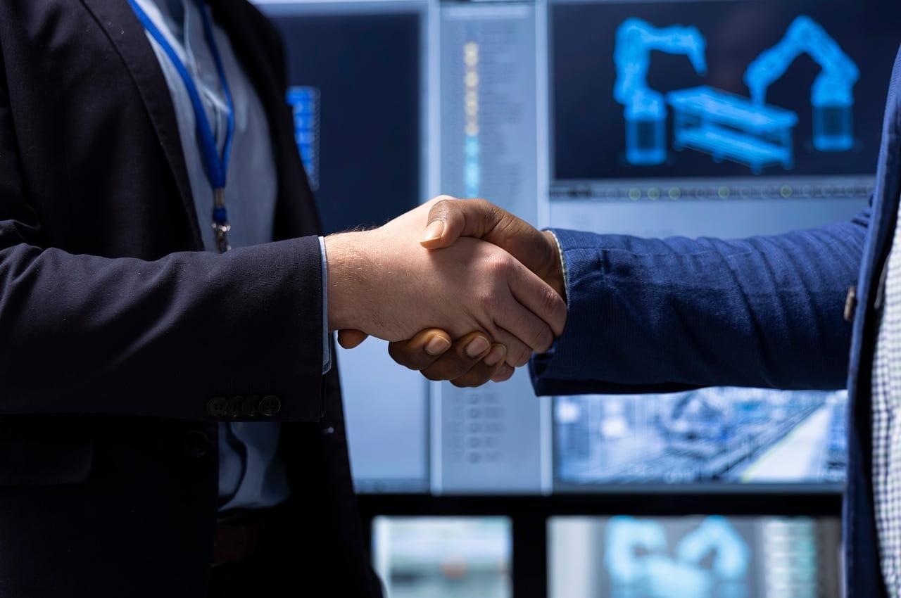 Homens apertando as mãos, com telas de computador ao fundo exibindo gráficos e dados, simbolizando um acordo ou fechamento de um negócio.
